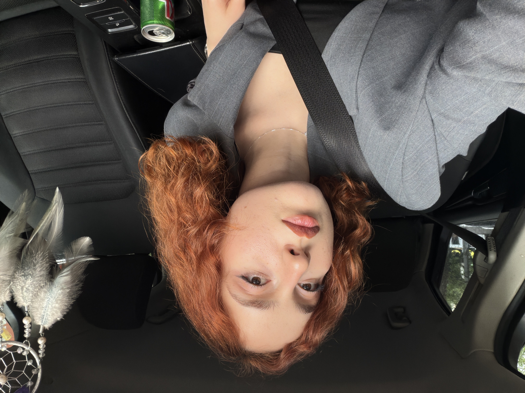 The image depicts a person with auburn curly hair inside a vehicle, wearing a gray blazer and a seatbelt. The camera is positioned overhead, resulting in an inverted perspective. An open can of green liquid is visible in the background near the vehicle's dashboard. A dreamcatcher hangs from the car's headrest. The setting is inferred to be in the city of Tbilisi, Georgia, based on the surroundings visible through the car window.