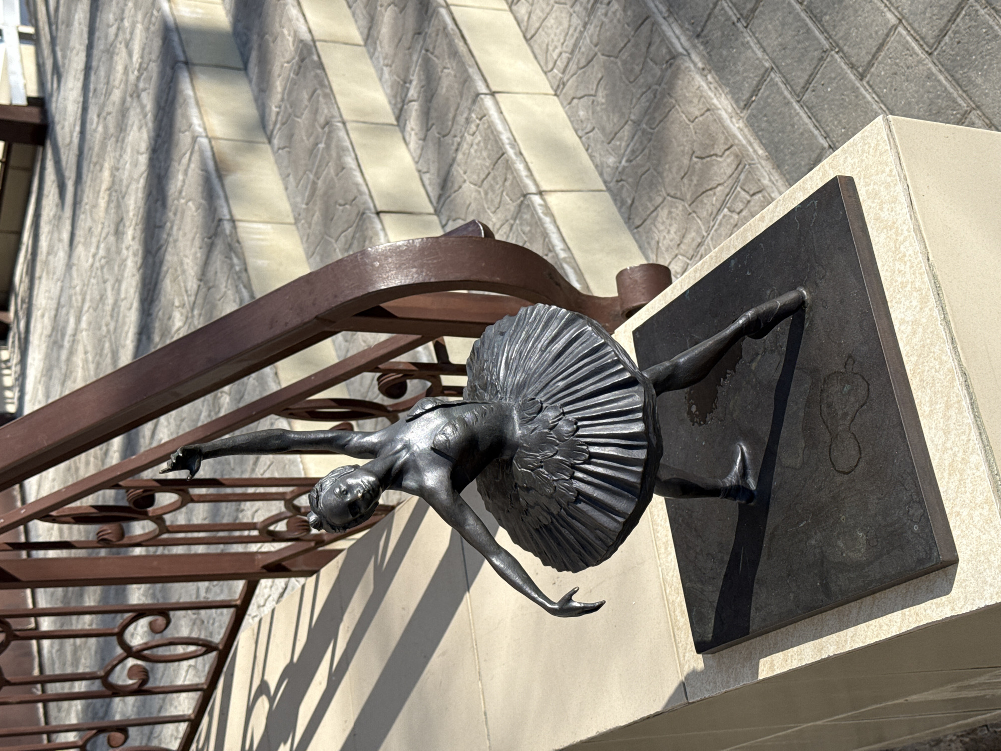 A dark-colored metal statue of a ballerina in a classical pose is positioned on a light beige tiled pedestal in Chaikovsky, Russia. The ballerina, wearing a multi-layered tutu, stands with her left arm extended vertically upwards and her right arm extended horizontally to the side, with fingers delicately curled. Her left leg is extended backwards on pointe, and her right leg is bent. The statue is mounted on a flat, dark rectangular base, which shows signs of light-colored residue or water marks.
Behind and to the right of the statue, a set of outdoor stairs ascends. The stair risers are light beige/cream, matching the pedestal, while the treads are textured grey paving stones with an irregular pattern. A dark brown metal railing with ornate scrollwork and vertical bars runs along the right side of the stairs. The scene is illuminated by bright sunlight from the upper left, casting distinct linear shadows from the railing onto the stairs and ground, as well as a shadow from the ballerina statue onto its base. The immediate foreground to the left of the pedestal consists of light beige tiles.