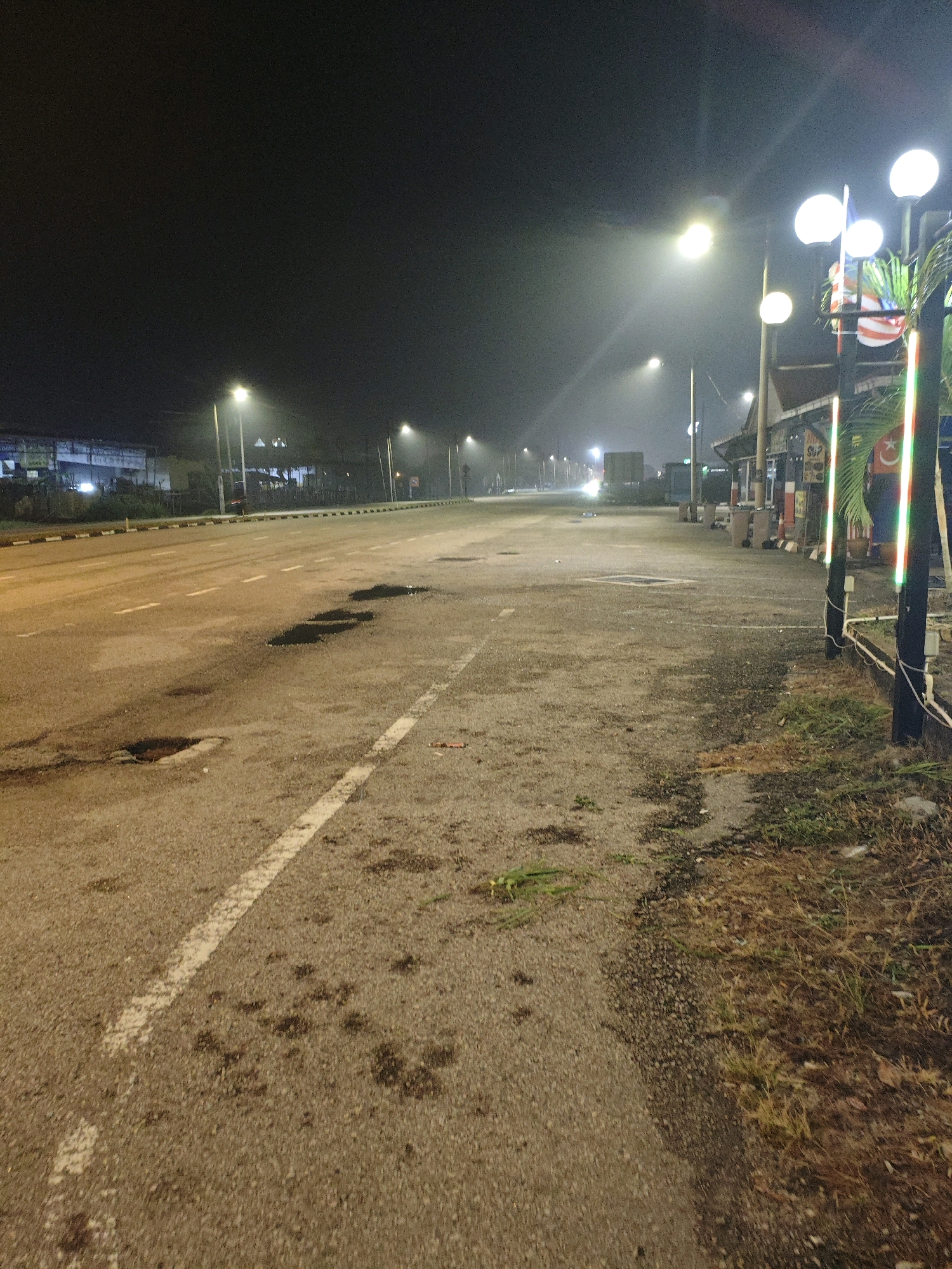 This photograph depicts a nighttime scene on a paved road in Kota Tinggi, Malaysia. The road appears to be a two-lane highway, with a white dashed line marking the center. Potholes are visible on the road surface, some filled with water. The surrounding environment is dimly lit, with streetlights casting a glow. On the right side of the road, there is a sidewalk or embankment with dry grass and some foliage. A building with a sign that reads "SUP" is visible in the background, along with decorative vertical lights. A Malaysian flag is also present, attached to a pole near the building. The sky is dark and there is a slight haze or fog in the air, contributing to the diffused lighting. There are no people or vehicles clearly visible in the immediate foreground or midground.
