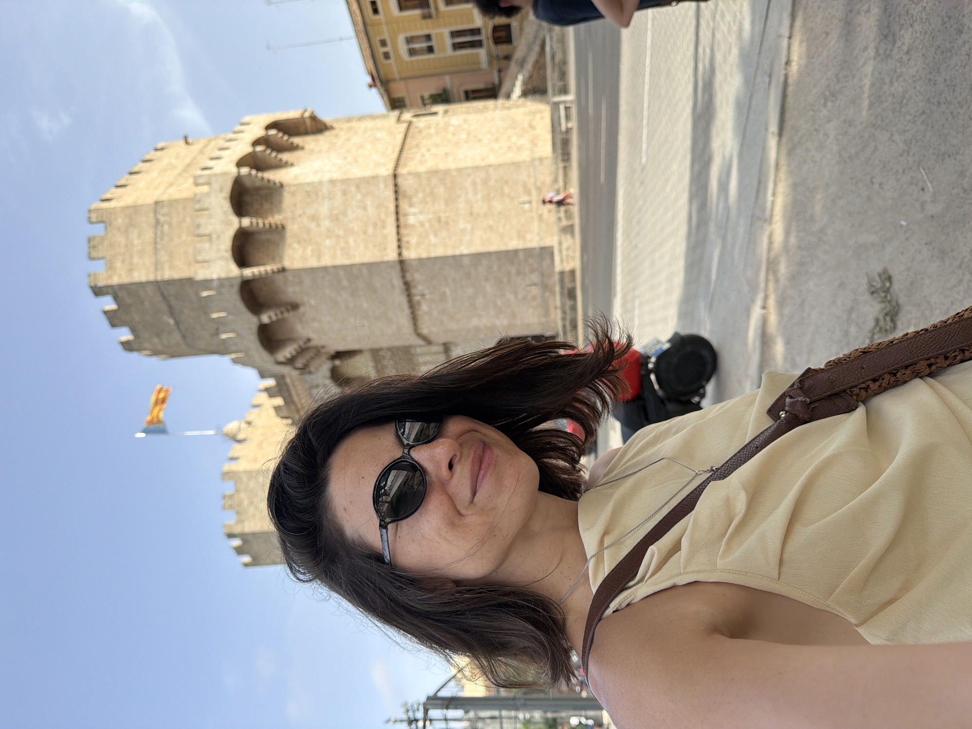 In a selfie taken outdoors, a woman with dark, shoulder-length hair and sunglasses smiles at the camera. She wears a light yellow, draped top and a brown crossbody bag with a woven strap. Behind her, a large, stone tower with crenellated battlements and arched windows dominates the background. A flag flies from the top of the tower. The setting appears to be a city square with a paved ground, a street, and buildings visible in the distance.