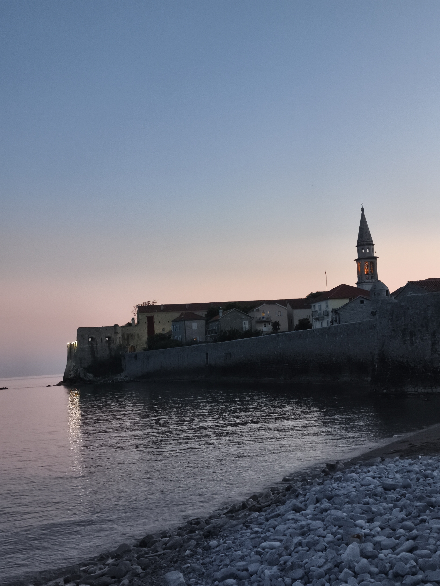 An atmospheric long shot captures the old town of Budva, Montenegro, at twilight or dawn. The historic fortified town is built on a rocky promontory, with rugged stone walls extending into the calm Adriatic Sea. The sky above transitions smoothly from a soft cerulean blue at the top to a delicate pink-orange hue near the horizon, indicative of either sunrise or sunset.
Artificial lights glow in several windows of the stone buildings within the old town and illuminate parts of the fortress walls, casting warm reflections onto the dark, placid water. A prominent bell tower with a distinct spire and cross rises above the other structures, its belfry also illuminated. The old town buildings feature traditional architecture with tiled roofs.
In the foreground, a pebbled beach slopes gently into the water, with individual smooth stones clearly visible. The water shows subtle ripples, reflecting the ambient light and the bright spots from the town. No people or active events are present, conveying a sense of timeless tranquility and historical grandeur. The weather appears clear and calm. There is no visible text.