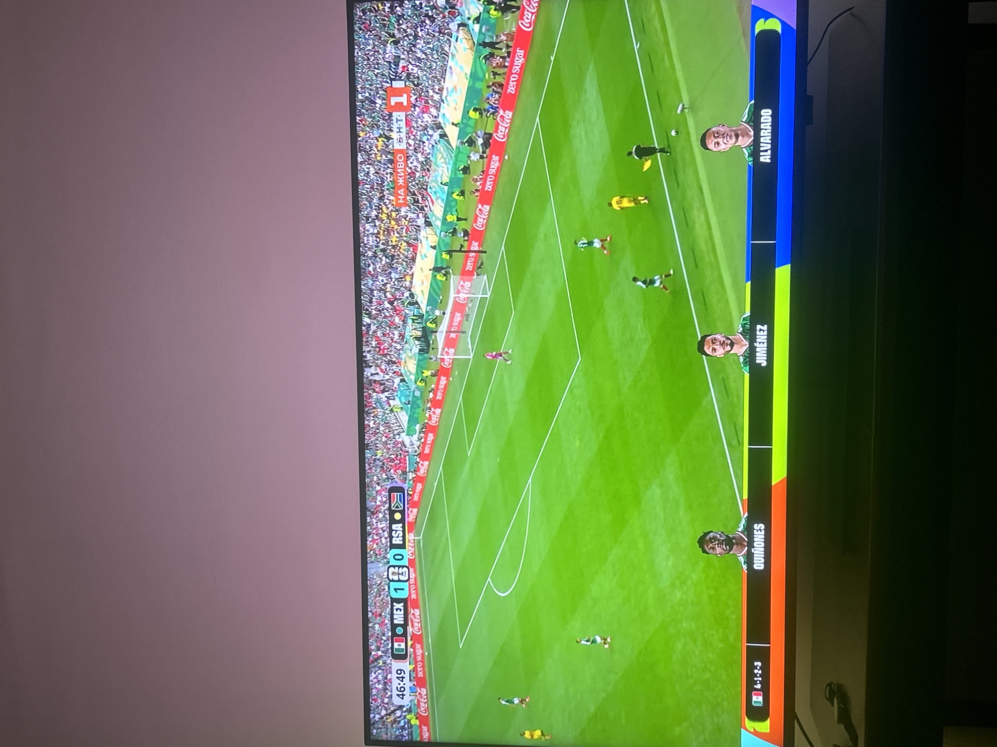 The image shows a live broadcast of a soccer match on a television screen. The score is displayed as 1-0 in favor of MEX (Mexico) against RSA (South Africa) at the 46:49 mark. The game is being broadcast live on BNT (Bulgarian National Television). The broadcast includes player information at the bottom of the screen, with "QUIÑONES", "JIMÉNEZ", and "ALVARADO" identified. The game is being played on a green grass field with white markings. A crowd is visible in the background, seated in stadium stands. The television screen is placed on a dark surface, likely furniture, with a pinkish-purple wall in the background above the screen. The scene is indoors, in a room with controlled lighting. There is no indication of the specific location within Pleven, Bulgaria, other than the broadcast being on Bulgarian National Television.