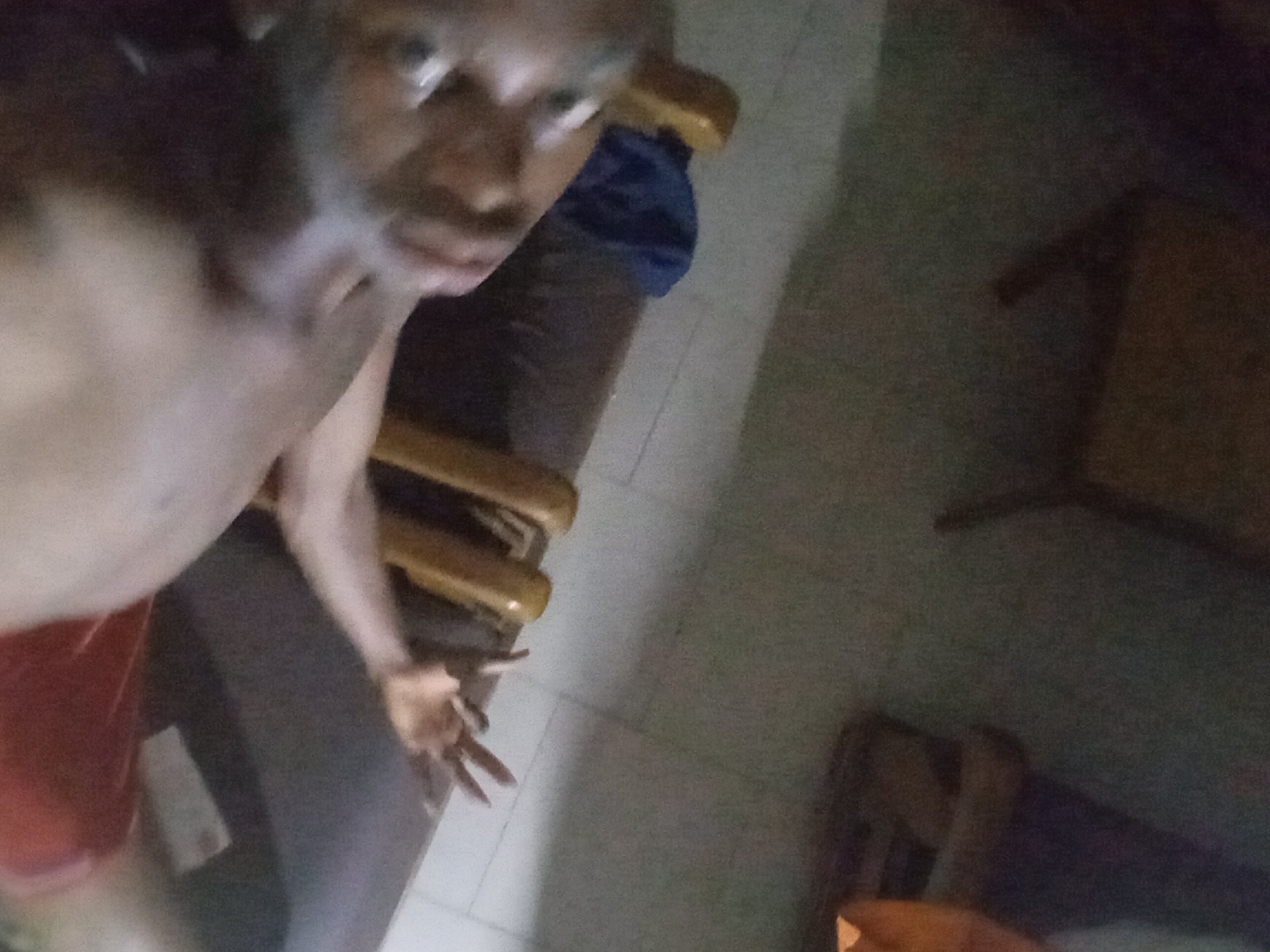 A dimly lit, close-up photograph of a shirtless dark-skinned person taken indoors. The person is facing the camera with an arm extended, and is wearing red shorts. Behind the person is a chair with a wooden frame and a blue cloth. The floor is tiled. Two wooden tables are visible in the background. The location is given as Nkpor, Nigeria.