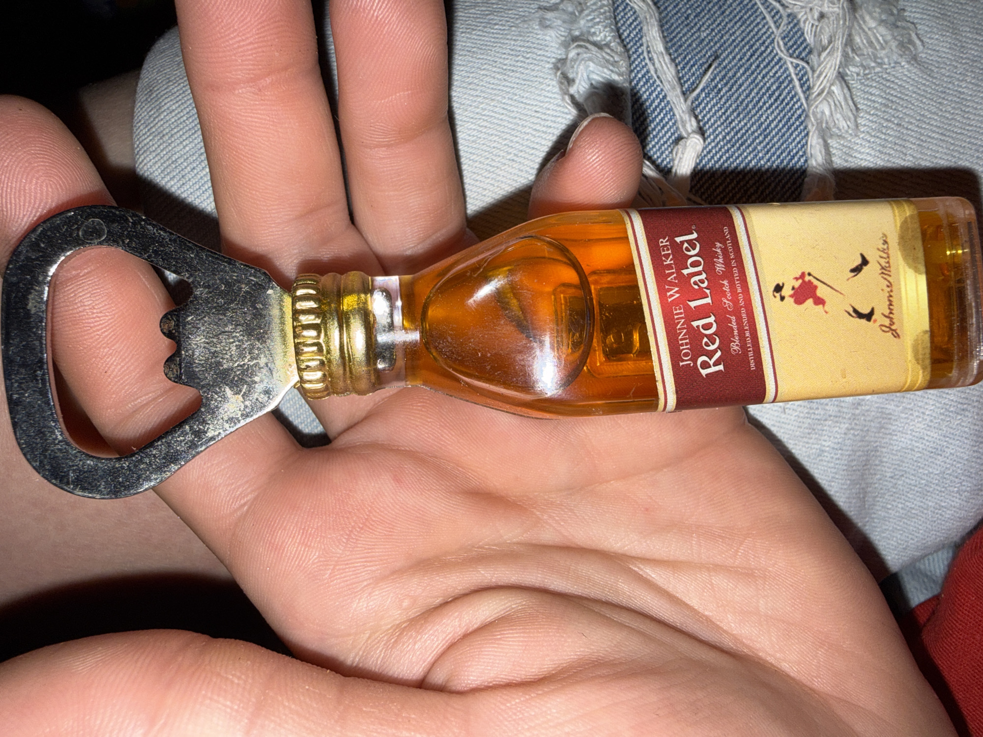 A human hand with fair skin tone is prominently featured, holding a miniature bottle opener designed as a replica of a Johnnie Walker Red Label whisky bottle. The fingers are curled around the bottle, revealing detailed skin texture and fingerprints. The bottle opener consists of a silver-colored metal head, showing minor scratches and wear, attached to a small, transparent bottle. The bottle contains an amber-colored liquid, and its label clearly reads "JOHNNIE WALKER Red Label Blended Scotch Whisky DISTILLED, BLENDED and BOTTLED IN SCOTLAND," accompanied by the "Striding Man" logo and "Johnnie Walker" signature. A gold-colored, ridged cap or collar connects the bottle to the metallic opener. The background is out of focus, displaying textile elements: light blue denim fabric, consistent with jeans and showing distressed areas with frayed threads, occupies the right side of the frame. A smaller portion of solid red fabric is visible in the lower-left corner. No specific architectural or environmental features are present in the image to indicate a geographic location, therefore the city of Stryama, Bulgaria cannot be confirmed.