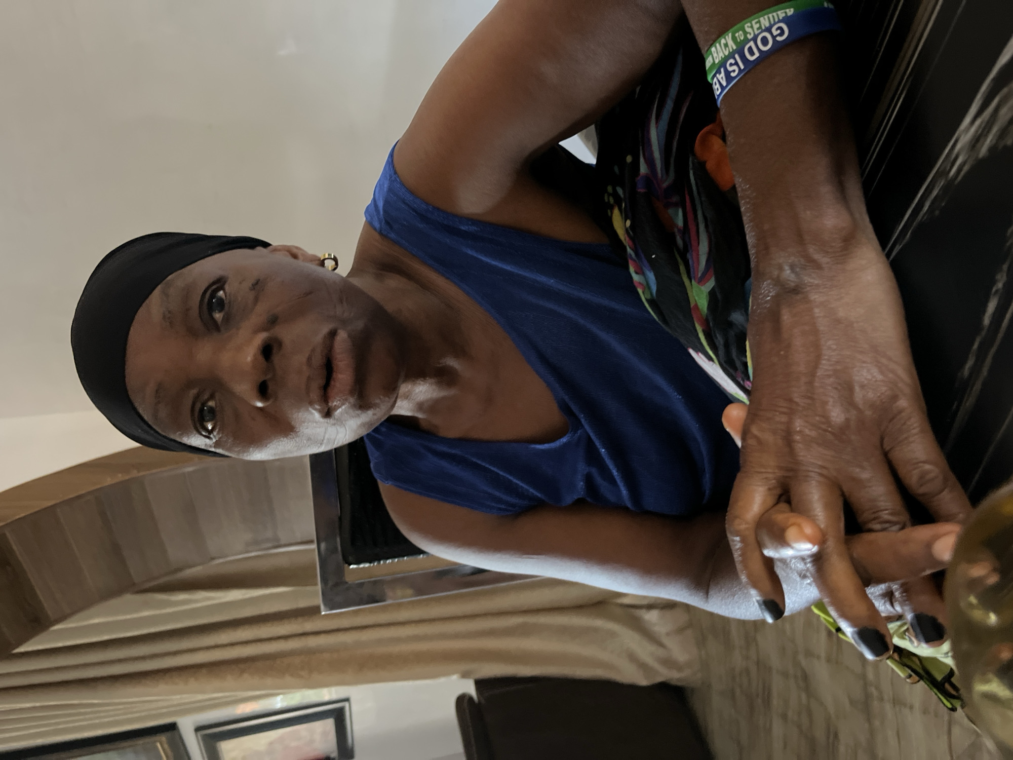 An older, dark-skinned woman with visible aging, wearing a black head covering and a blue tank top, is sitting indoors. She has a green and blue wristband that says "GOD IS ABLE" on her right wrist. Her hands are clasped together, showing dark nail polish on some of her nails. The background includes beige curtains and a framed picture on a wall. A chair is visible. There is no evidence in the image to indicate the location is Jos, Nigeria.