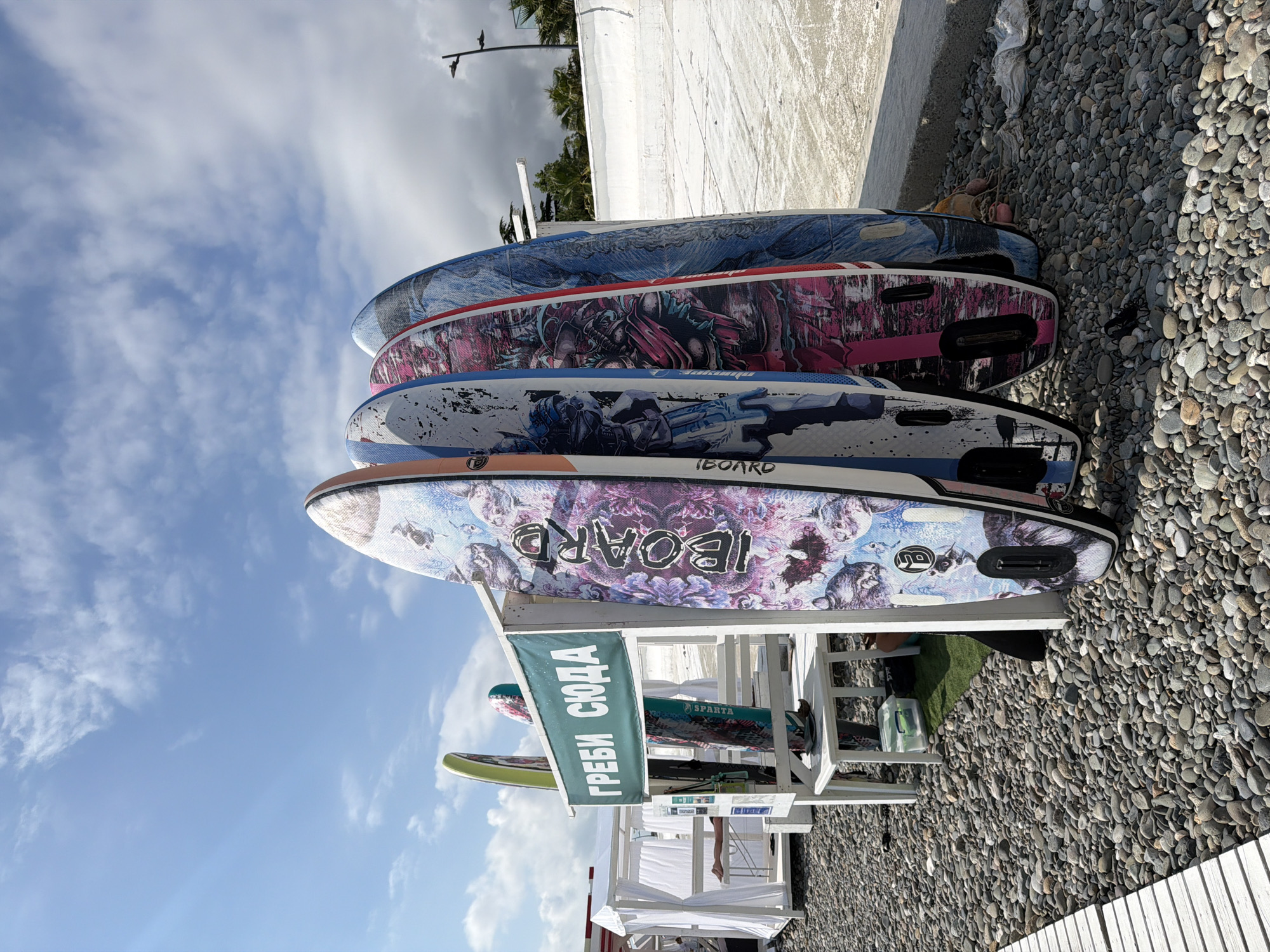 In Sirius, Russia, on a pebble beach, several stand-up paddleboards are leaning against a white wooden structure that displays a green banner with the Russian text "ГРЕБИ СЮДА," meaning "Paddle Here." The paddleboards have vibrant, intricate designs. To the left of the structure, a white cabana with draped curtains is visible. The scene is set against a backdrop of a bright blue sky with scattered clouds, a white wall, and some palm trees in the distance. The ground is covered in small, smooth pebbles.