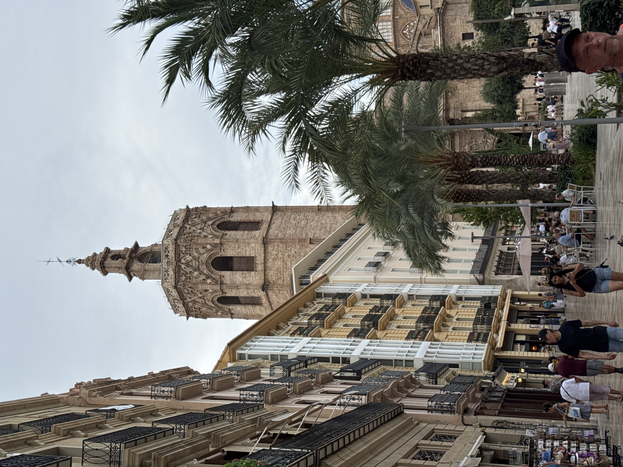 The image captures a view of Valencia Cathedral's Miguelete Tower, a prominent landmark in Valencia, Spain, from a street lined with buildings and palm trees. The Miguelete Tower, a Gothic bell tower, is visible in the upper center of the frame, characterized by its octagonal base and intricately carved stone details. To the left, a row of multi-story buildings with balconies and various architectural styles forms the streetscape. Some buildings feature balconies with wrought-iron railings, while others have modern glass window installations.
In the foreground and mid-ground, several palm trees are prominent, their fronds reaching towards the overcast sky. The street itself appears to be a pedestrian walkway, paved with light-colored stones. People are scattered throughout the scene, some walking along the street, others seated at outdoor cafes or browsing merchandise displayed on racks. In the immediate foreground, a man wearing a black baseball cap is visible, looking towards the camera. The overall atmosphere suggests a public space in a European city, likely a popular tourist area given the presence of shops and cafes. The sky is cloudy, indicating overcast weather.