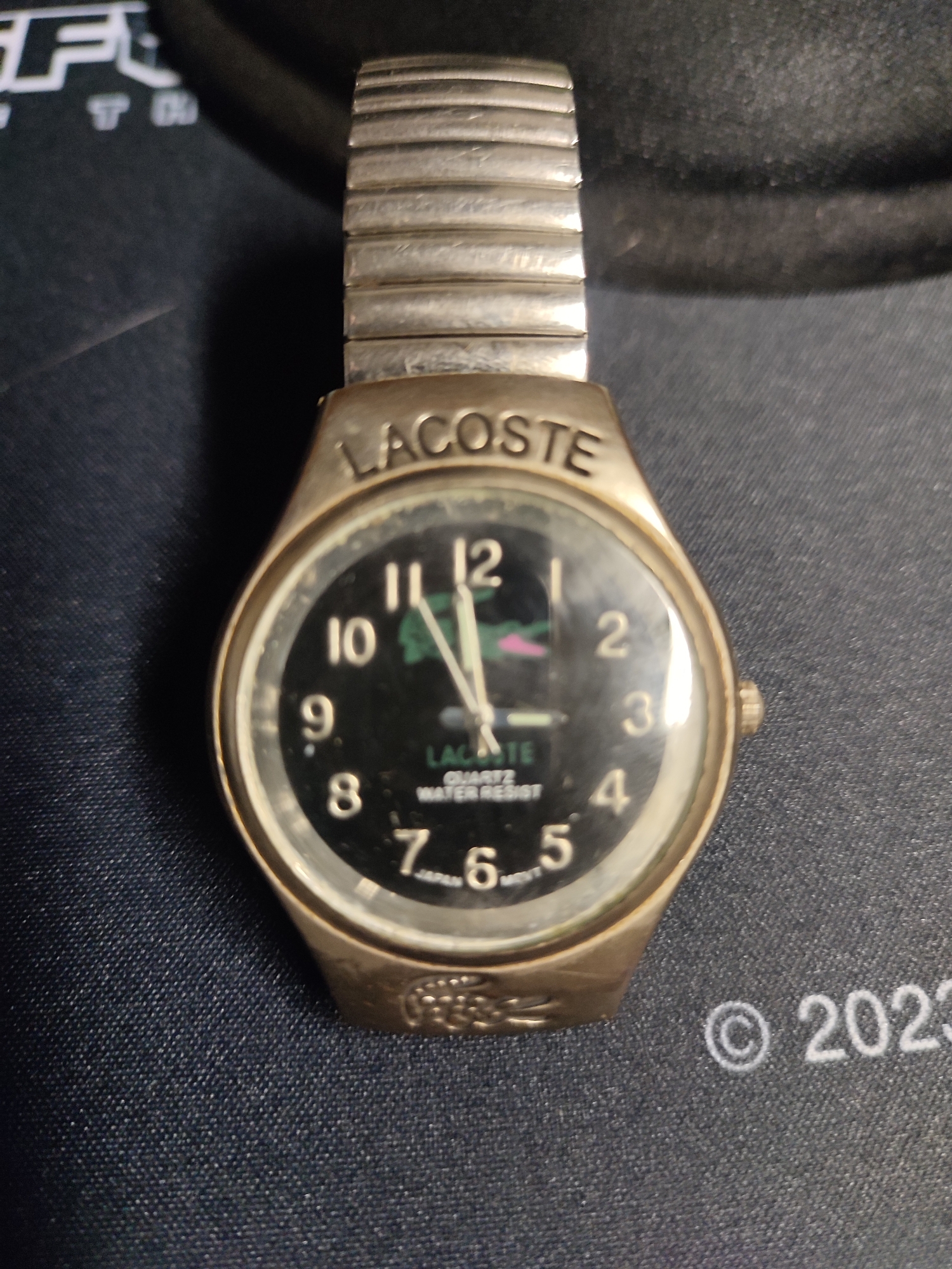 A close-up, top-down shot of a gold-toned, metal-banded wristwatch with a black face is positioned on a dark blue, textured surface. The watch features silver hour markers and hands, with the brand name "LACOSTE" clearly visible above the face and "QUARTZ WATER RESIST" and "JAPAN MOVT" printed on the dial. The watch band is segmented and metallic. To the right of the watch, the year "2023" is visible, also printed on the dark surface. The background is dark and out of focus, but hints of lettering, possibly from a logo, are present in the top left corner. There is no indication of the location being Biliakiemis, Lithuania.