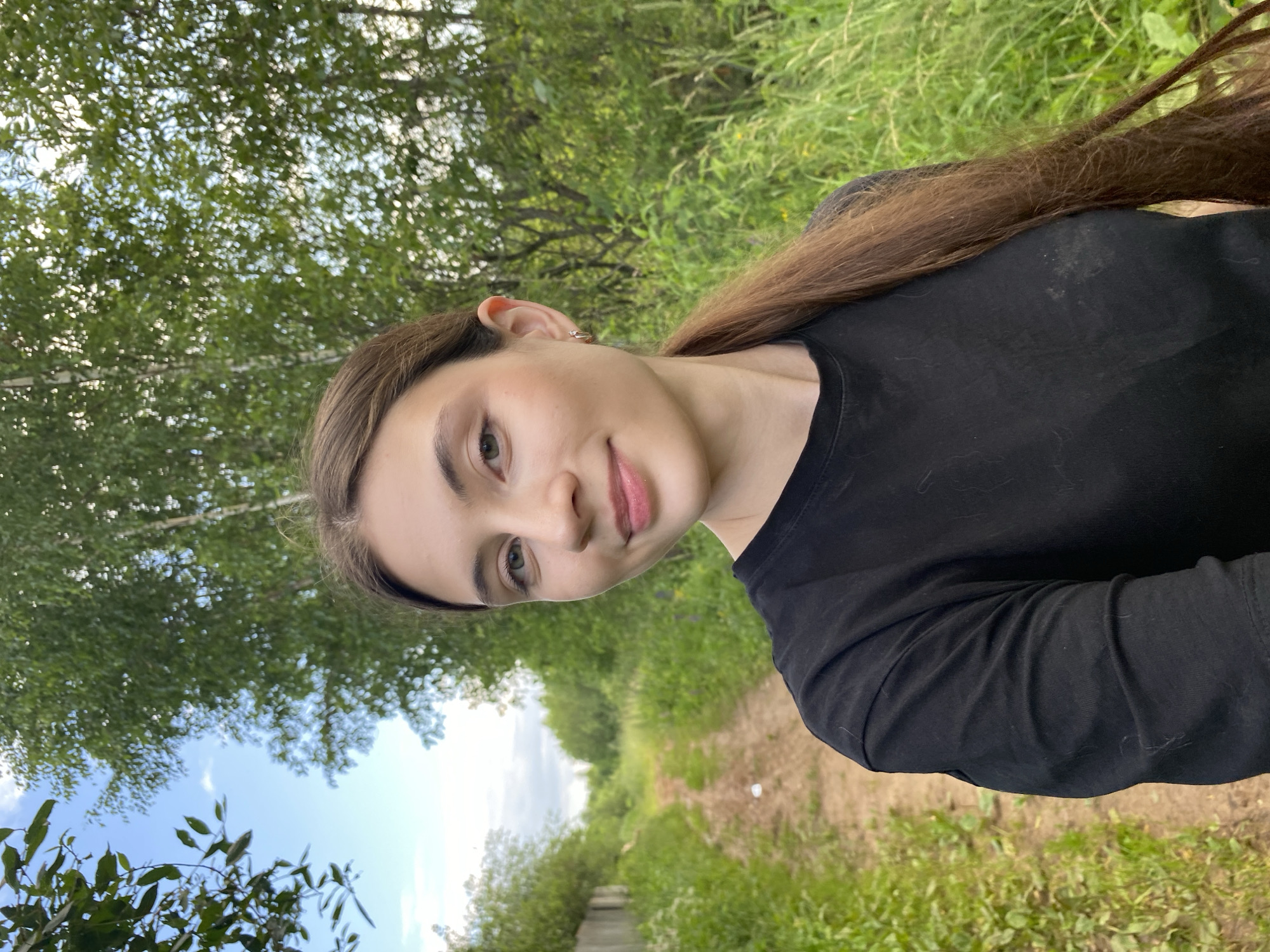 The media file is a portrait of a young woman outdoors. She is facing towards the camera, with her head tilted slightly. She has long, brown hair that is partially visible. Her eyes are open, and she is smiling subtly. She is wearing a black long-sleeved shirt.
The background consists of lush green foliage, suggesting a natural setting like a park or a forest. A dirt path or road is visible in the distance, winding through the greenery. The sky is partly cloudy with patches of blue. The lighting suggests it is daytime, likely late afternoon, with soft, natural light.
The overall mood is relaxed and peaceful. The woman's expression conveys a sense of contentment. There are no visible objects or text in the image. The scene appears to be set in a rural or semi-rural area, consistent with the provided location context of Novyi Ural, Russia. The image is oriented vertically, as if taken with a phone.