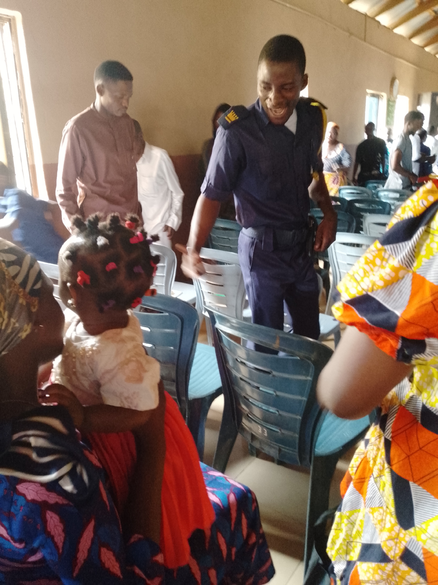 The image captures a scene indoors, possibly within a community hall or a church-like setting. A man in a navy blue uniform with shoulder markings and a belt is interacting with people. He has a light complexion and is smiling, suggesting a friendly interaction. A woman seated is holding a young child with intricately styled hair adorned with colorful decorations. The child is facing away from the camera. To the right of the frame, another woman is seen wearing a vibrant, patterned garment. A stack of blue plastic chairs is arranged in the background. The environment seems to be in the city of Unknown, Unknown. There are multiple other people in the background, contributing to a sense of an event or gathering. The room has natural light coming in from windows, and the walls appear to be painted in neutral tones.