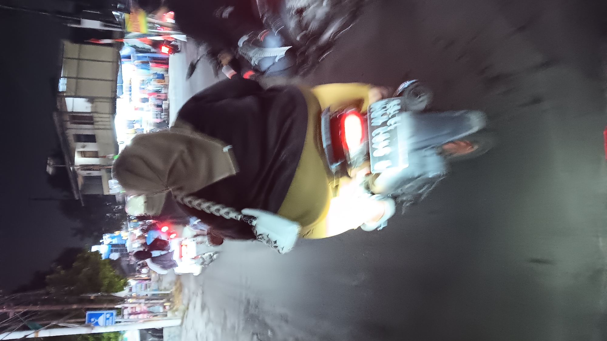 The image captures a night scene on a street in Depok, Indonesia, likely near a commercial area with shops and stalls lining the road. The primary focus is on a person riding a motorcycle, with the camera positioned behind and slightly above them. The rider is wearing a hijab and a dark top, with bright yellow pants visible. They are carrying a light-colored shoulder bag with a distinctive braided strap. Another motorcycle with a rider in a white helmet is visible ahead, also moving in the same direction. The street is dark, illuminated by streetlights and the lights from the shops. Some buildings are visible in the background, appearing to be two-story structures with awnings over the shopfronts. There are other vehicles and pedestrians in the background, but they are blurred due to motion or low light conditions. The overall atmosphere suggests a busy, active street at night.