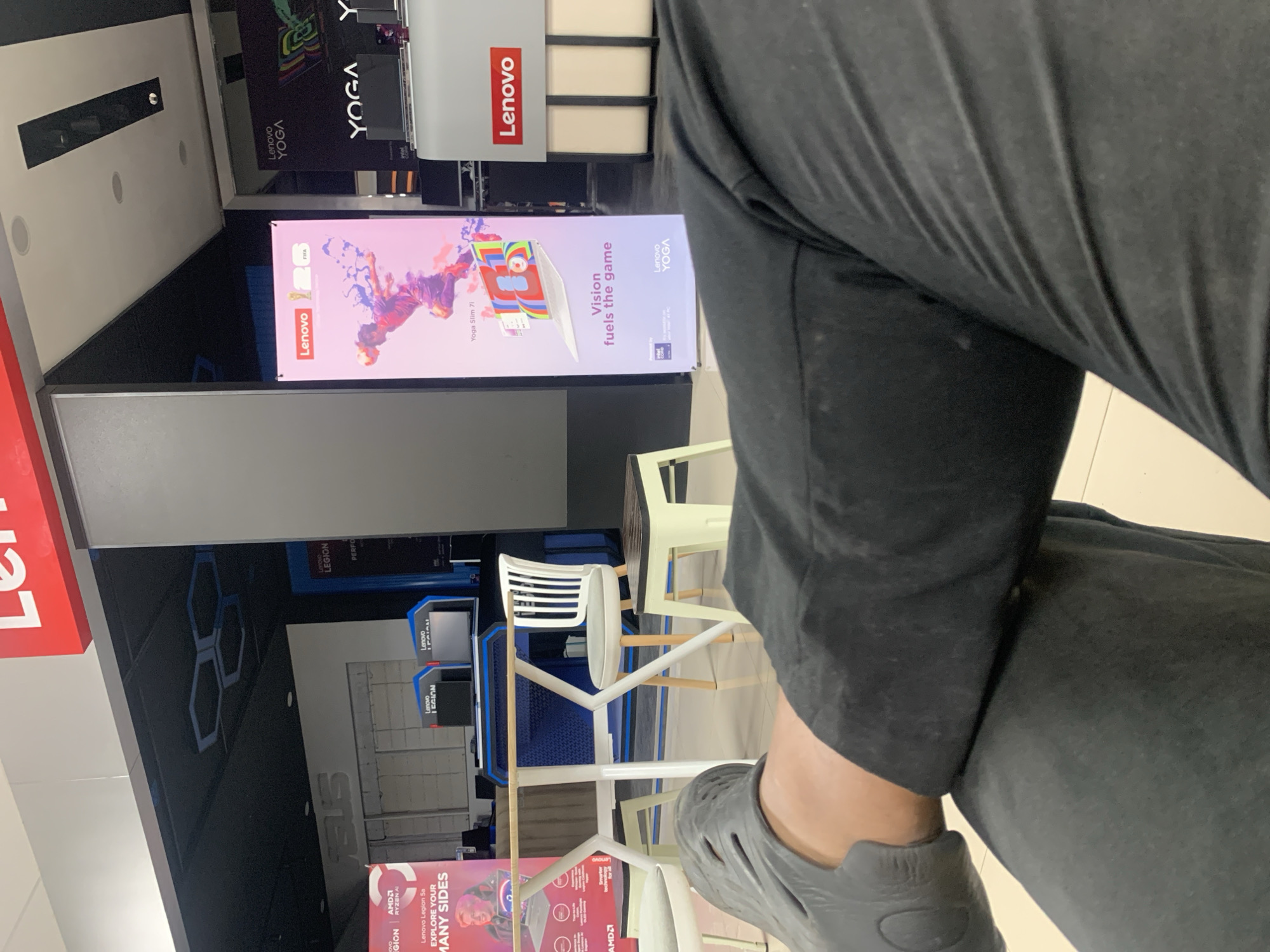 The image captures an indoor scene, likely a retail store or showroom, with prominent branding for Lenovo and ASUS. In the foreground, a person's legs and feet, clad in dark pants and gray sandals, are positioned across the frame, obscuring much of the lower portion of the scene.
The background reveals a display area with various Lenovo laptops and promotional materials. A large vertical banner showcases a Lenovo Yoga Slim 7i laptop with the tagline "Vision fuels the game," featuring colorful abstract graphics and an image of a soccer player. Another display features the "Lenovo YOGA" branding. Two Lenovo Legion monitors are visible on a blue and black display stand. A white table with a light-colored wooden top is centrally placed, surrounded by chairs, including white plastic chairs with wooden legs and a light-colored wooden stool.
Behind these displays, a large white ASUS logo is visible on a wall, suggesting a co-branded or multi-brand retail space. The ceiling features dark panels with hexagonal light fixtures and recessed lighting. The overall environment appears to be a well-lit retail space. The text "Palembang, Indonesia" is not visible in the image and cannot be confirmed.