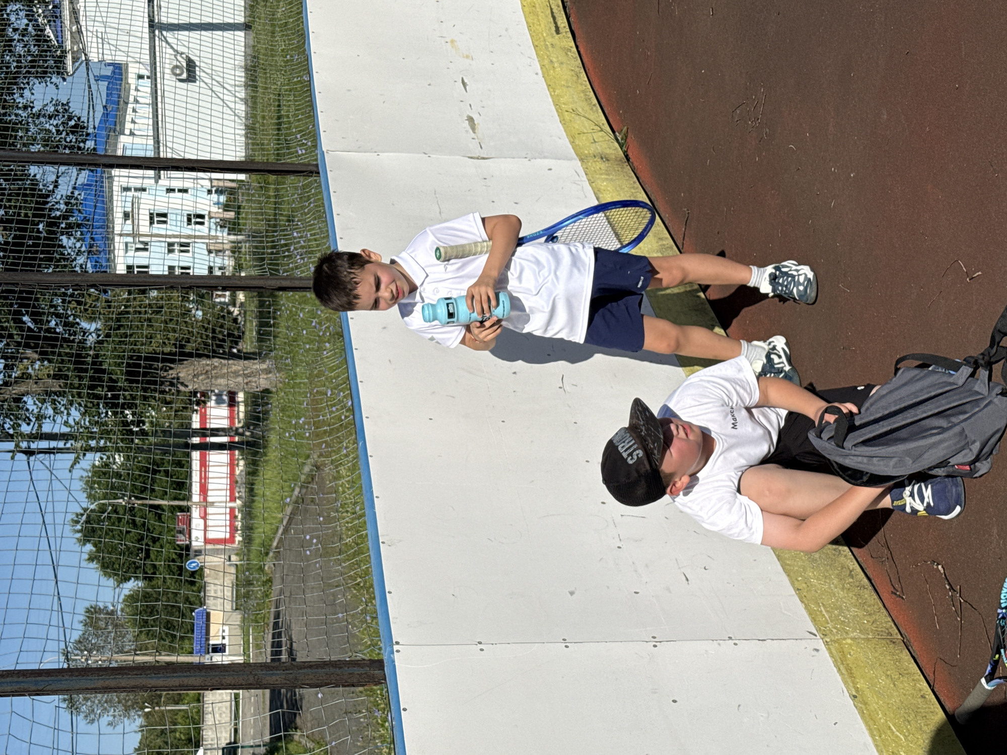 Two young boys are on an outdoor sports court, possibly a tennis or basketball court, in Chaikovsky, Russia. The court surface is reddish-brown. A white barrier with a blue stripe runs along the edge of the court. In the background, a chain-link fence separates the court from a grassy area with trees and buildings. One boy, wearing a white polo shirt and navy blue shorts, stands holding a blue water bottle and a tennis racket. He is looking down and to his left. The other boy, wearing a white t-shirt, black shorts, and a black baseball cap, is crouched on the ground. He is looking to his right and holding a dark gray backpack on his lap. His t-shirt has "Marcus" written on it. Both boys are wearing athletic shoes. The sun is shining, casting shadows on the court.