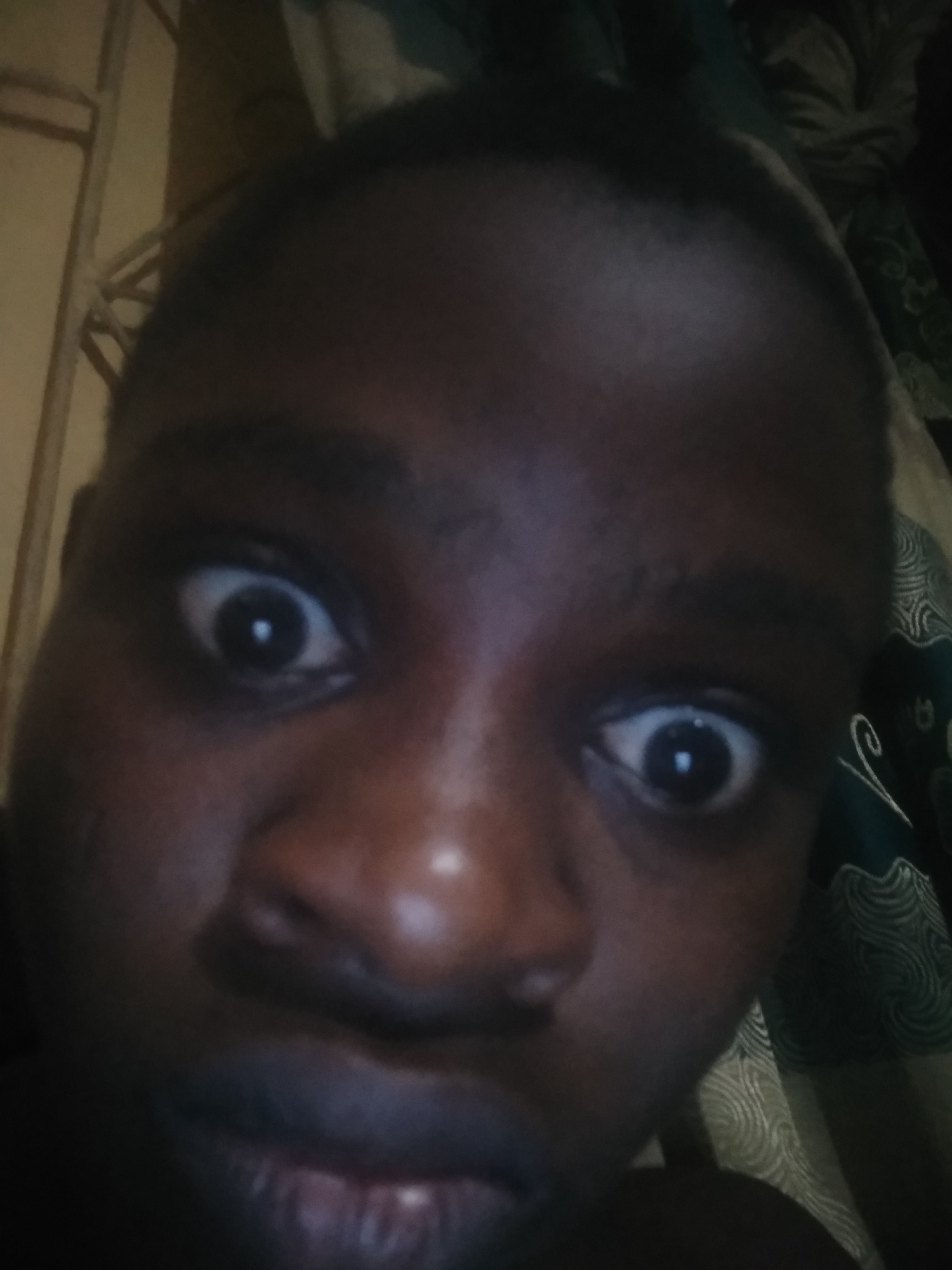 A close-up, selfie-style portrait features a person with dark skin, whose face fills most of the frame. The individual looks directly at the camera with wide-open eyes, showcasing dilated pupils and distinct light reflections, conveying an expression of surprise, shock, or playful exaggeration. Their mouth is slightly agape or in a relaxed, slightly pouted position. Short, dark hair is visible on top of the head.
The background is dimly lit, suggesting an indoor setting. On the left side of the frame, a vertical light-colored structure, possibly a door frame or wooden panel, is partially visible. On the right, a patterned fabric, likely a curtain or bedding, features a green or teal color with intricate white or light grey swirling designs. No text is visible in the image.
