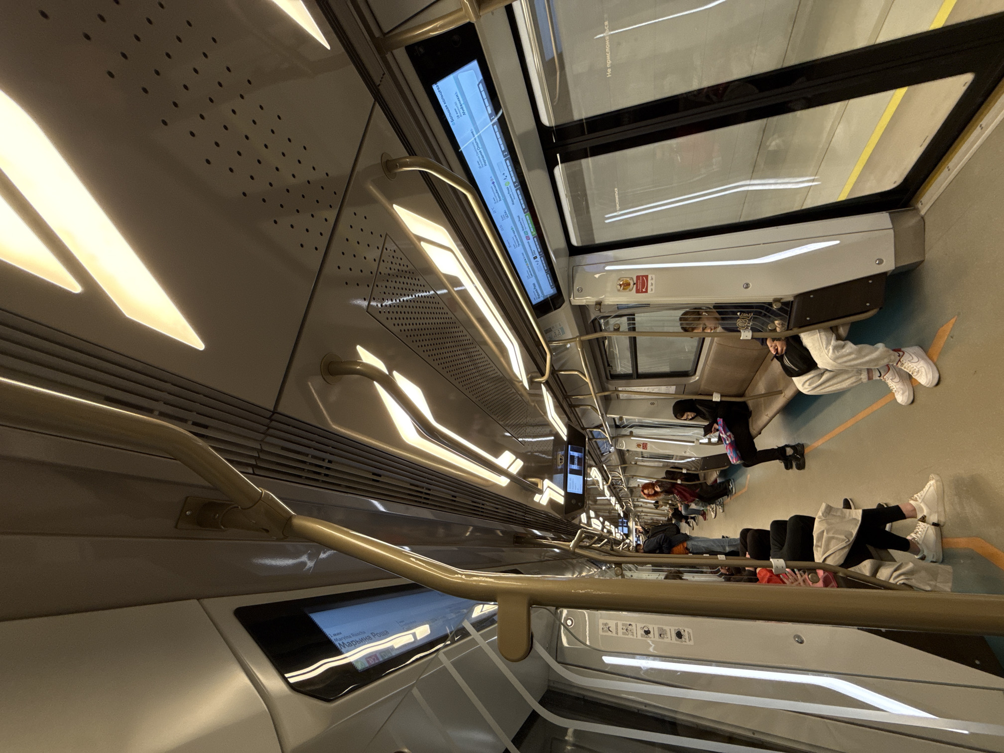 The image displays the interior of a modern subway train car, likely in Moscow, Russia, as evidenced by the Cyrillic text on the digital displays and station names. The perspective looks down the brightly lit aisle, showcasing the train's contemporary design. Overhead, warm, rectangular lights illuminate the sleek, light-colored ceiling and walls, which incorporate perforated panels. Brass-colored handrails extend along the ceiling and sides of the car, providing support for standing passengers.
Multiple digital display screens show route information in both Cyrillic and Latin scripts. Visible station names include "Марьина Роща" (Maryina Roscha), "Мневники" (Mnyovniki), and "Народное Ополче" (Narodnoye Opolcheniye), all associated with Moscow's "Большая кольцева" (Big Circle Line). One screen also displays "1 мин," indicating a one-minute countdown.
Several passengers are visible, mostly seated, engaged in typical commuting activities. Closest to the camera, a person wearing light-colored sweatpants, white sneakers, and a face mask sits with a dark bag across their chest. Further down the aisle, other commuters are present, some also masked. Through the windows, reflections and blurred glimpses of what appears to be a subway platform or tunnel can be seen. The text "Не прислон" (short for "Do not lean") is visible on a window panel. The scene conveys a calm, everyday commuting environment. The time of day and weather are not discernible from the interior view.