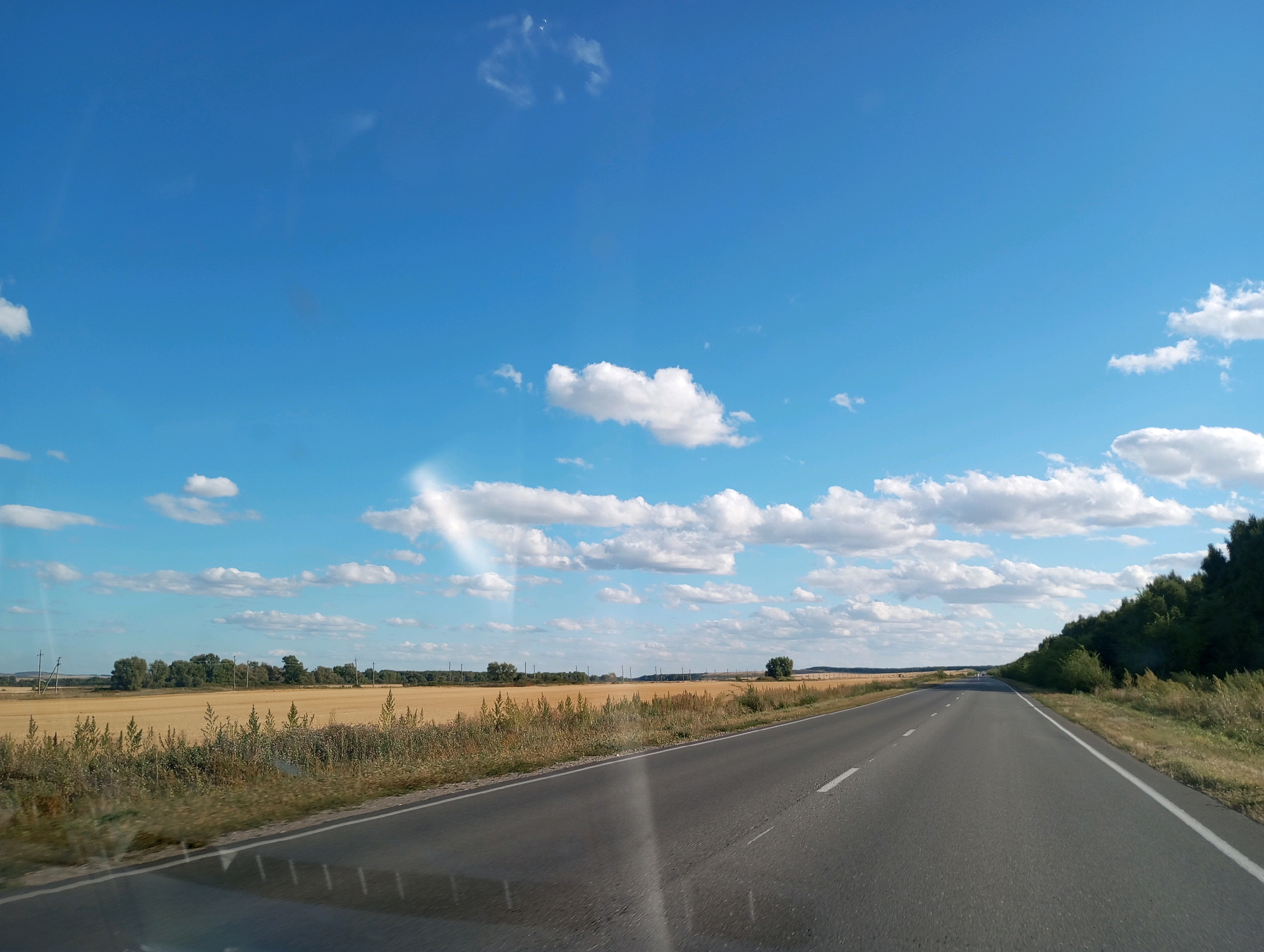 The image shows a paved road stretching into the distance, bordered by dry grass and fields of harvested crops. The sky is a vibrant blue with scattered white clouds, and a light sunbeam is visible on the right side of the frame. A line of trees forms a dark green border along the right side of the road, and in the distance, there are more trees and what appear to be utility poles. The overall scene depicts a rural landscape, likely in Syrtinskii, Russia, on a clear day.