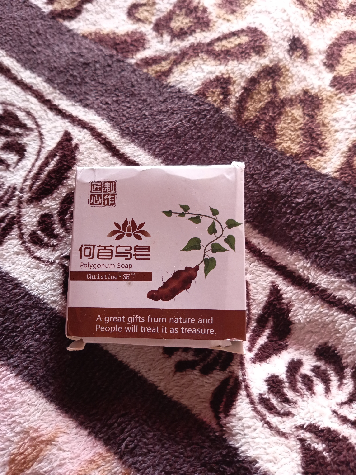 A square, light pink soap box is centered on a brown and white patterned fuzzy blanket. The box has Chinese characters on the top left, a lotus-like symbol below them, and then the words "何首乌皂" in larger Chinese characters. Beneath that, "Polygonum Soap" is written in English. Below that is "Christine SH TM" and a drawing of a plant with a root. At the bottom of the box is text that reads, "A great gifts from nature and People will treat it as treasure." The blanket has a leopard-like pattern. The lighting suggests it may be indoors, with no indication of a specific city or location.