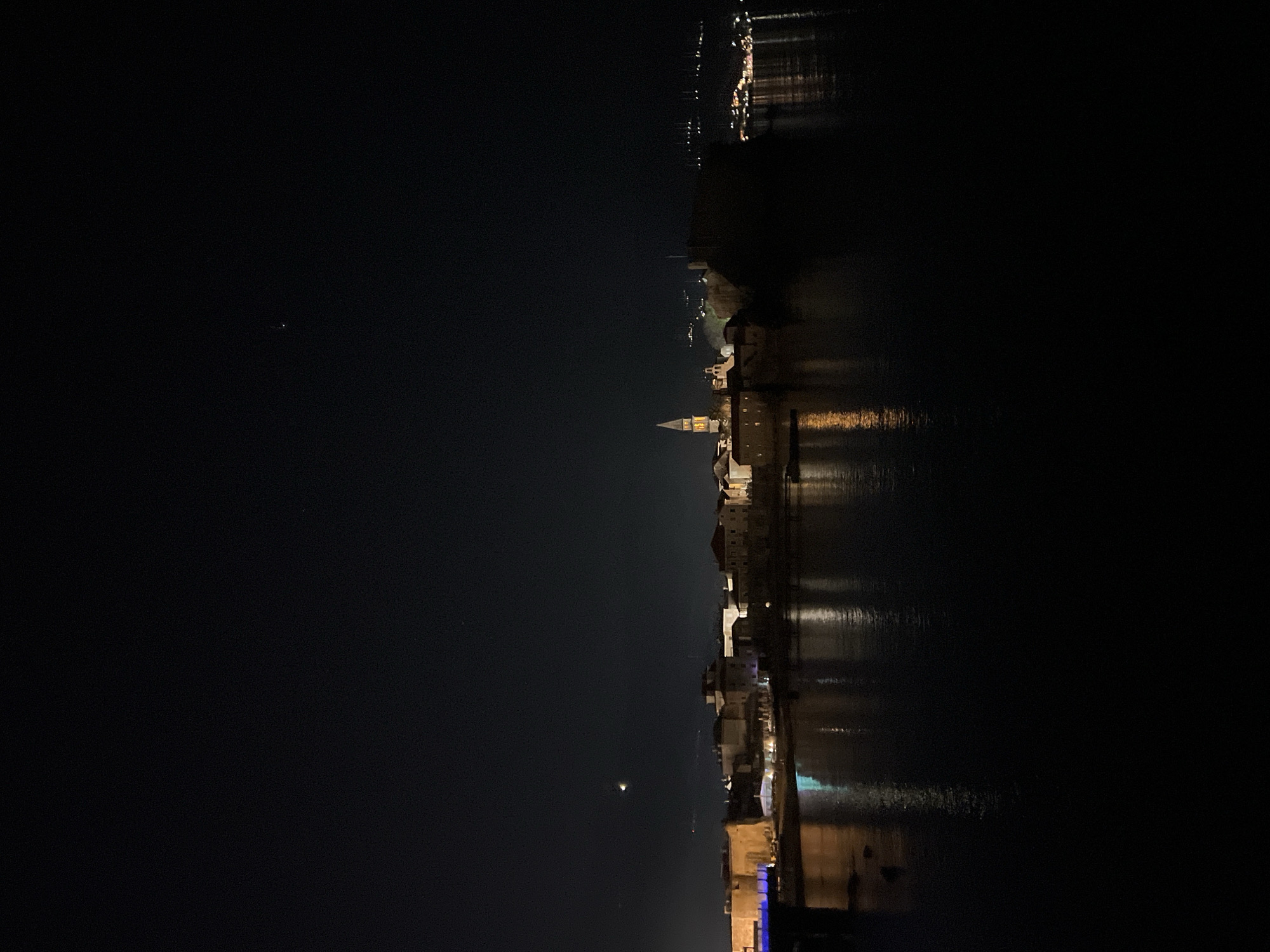 The image depicts a night scene of Budva, Montenegro, with a view of the Old Town skyline from across the water. The image is oriented vertically. The water reflects the lights of the Old Town, creating a mirrored effect. A few stars or planets are visible in the dark sky. The Old Town's notable structures, including the bell tower of St. John's Church, are illuminated. Other buildings closer to the viewer are partially lit with cool toned lights. The horizon appears to have other towns or settlements with dimly lit buildings. The surrounding environment is dark, emphasizing the lights of the town.