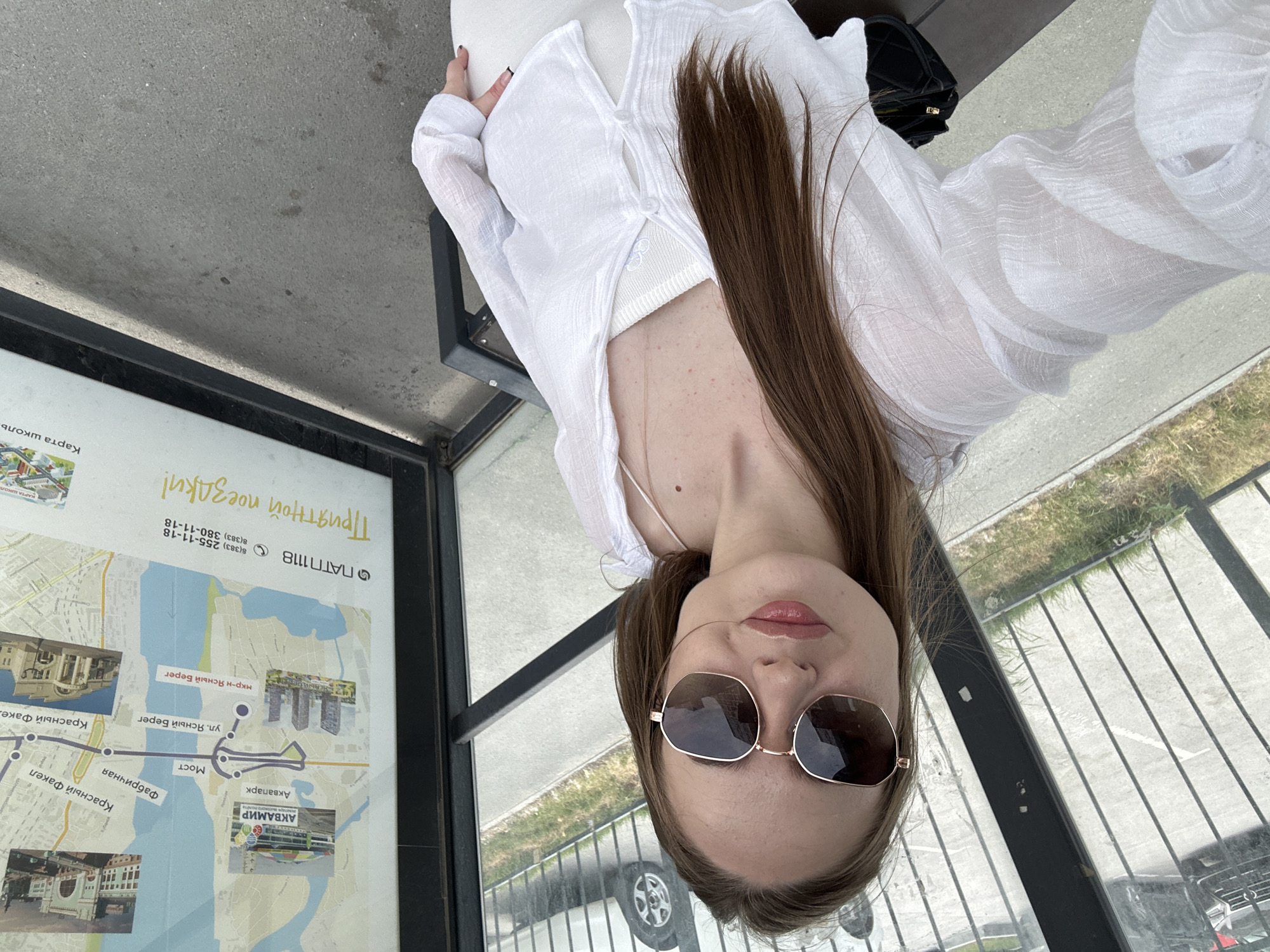 A young woman with long, brown hair, wearing oversized sunglasses, a white linen shirt, and a white tank top, poses for a selfie. She is standing in a bus shelter with a map displayed on the wall behind her. The map shows a city with various landmarks and street names. The floor of the bus shelter is concrete, and the area outside appears to be a street with parked cars and a grassy area.