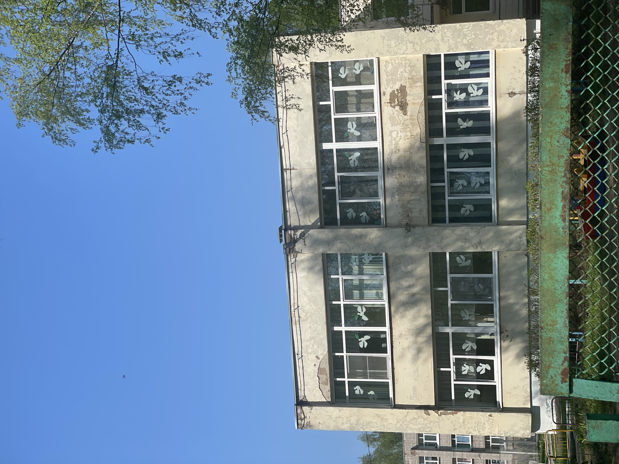 The image shows a building in Izhevsk, Russia, likely a school or kindergarten, identifiable by the white dove stickers on the windows. The building has a faded, aged facade, suggesting it's an older structure. A green chain-link fence is partially visible on the right side of the frame. Above the building, branches of trees are visible against a clear blue sky. Another building can be seen in the lower corner of the frame.