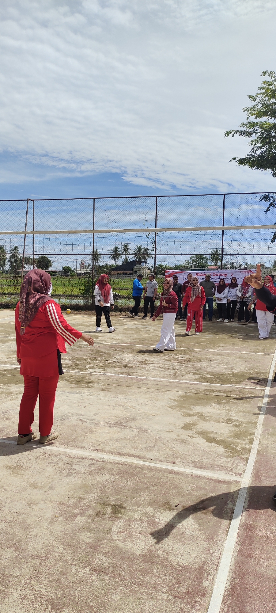 This is a full-frame, eye-level, outdoor shot of a group of women playing volleyball on a concrete court. The sky is blue with scattered white clouds. The court has white lines marking its boundaries. A volleyball net spans the middle of the court. In the background, there is a fence, some greenery, palm trees, and buildings. Some women are actively playing the game, while others are observing from the sidelines. The women are wearing a mix of red and white athletic attire, and most are wearing headscarves. The scene appears to be in Peurupok, Indonesia, based on the presence of palm trees and the general architectural style of the buildings in the background.