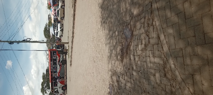 The image shows a brick paved street in Kitengela, Kenya, located at coordinates -1.47 latitude and 36.96 longitude. The street is empty except for a red and white bus that is parked on the left side of the frame. The bus has a sign that says "FISA TOUR" on the side. There are several people standing around the bus, but their faces are not visible. There are also several cars parked in the background, including a white minivan. The sky is blue with some white clouds. The pavement is shaded by the bus and a tree on the left. The image appears to be taken from a low angle, looking towards the street from the curb.