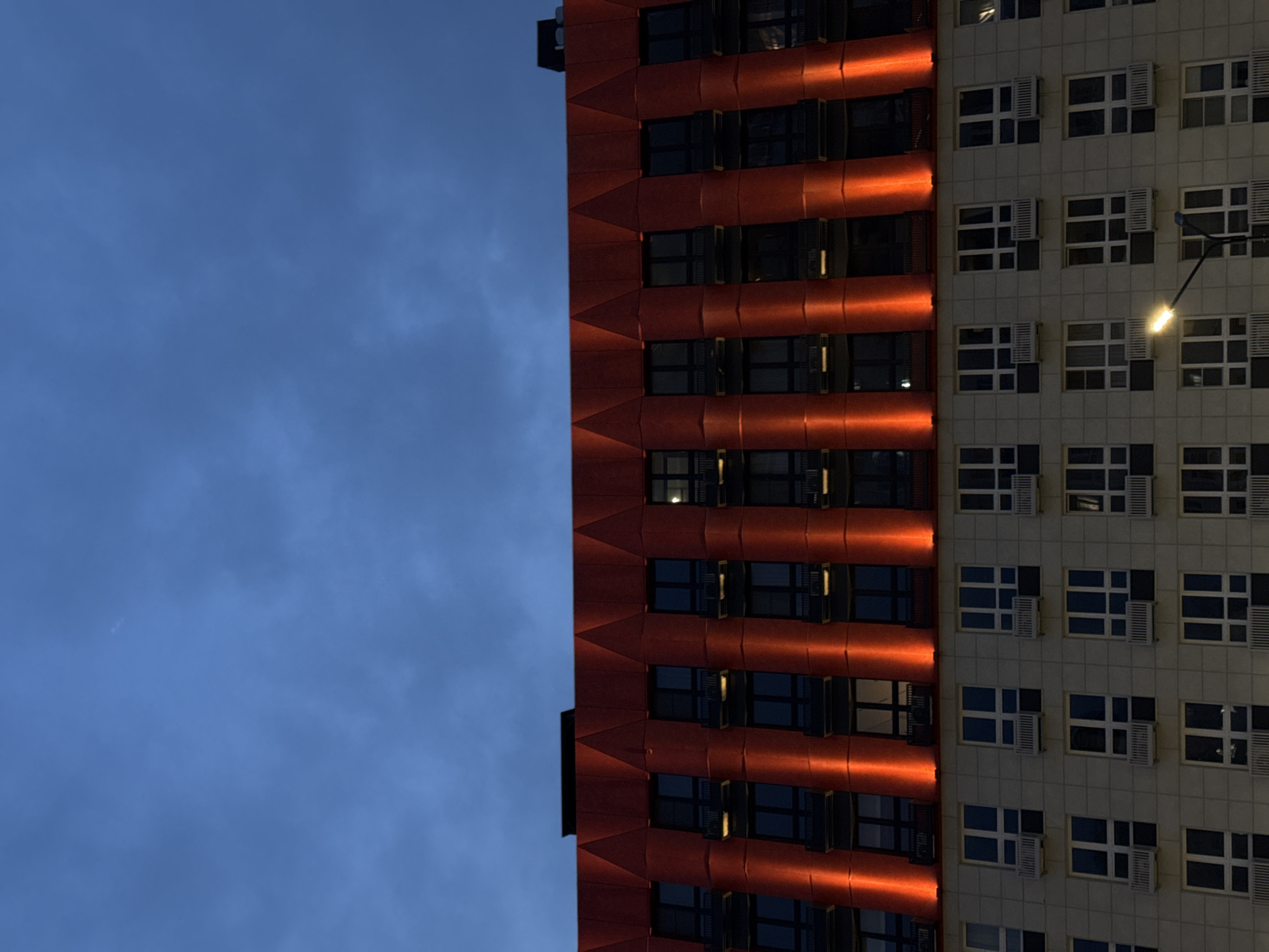 A low-angle, medium shot reveals a portion of a tall building under a blue, cloudy sky. The building's facade is divided into two distinct sections. The left portion of the facade is a burnt orange color, marked by horizontal ridges, and interspersed with dark windows. The right portion of the facade is grey and features a grid-like arrangement of windows, many with air conditioning units. A streetlight is visible on the right. The building is located in Nizhny Novgorod, Russia.