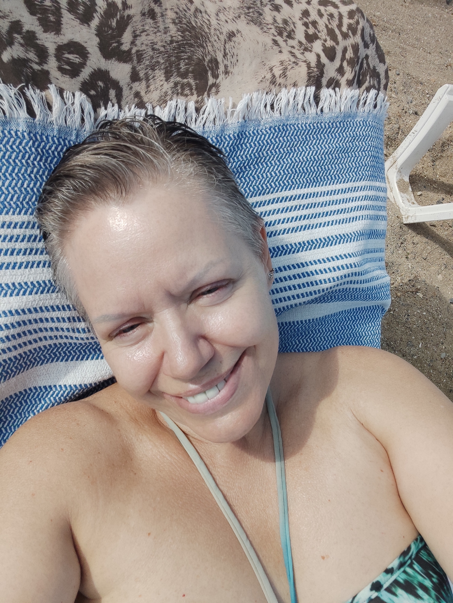 The media file shows a close-up, overhead shot of a woman lying on a beach chair covered with a blue and white striped towel. A leopard-print blanket is visible behind her head. The woman has short, graying hair, and she is smiling and looking towards the camera. Her skin appears slightly shiny, possibly from sweat or sunscreen. She is wearing a bikini top with a teal and green tropical pattern. The background shows sand and the edge of a white beach chair, suggesting an outdoor setting. The lighting indicates it is daytime, likely sunny given the beach environment. The woman's expression conveys happiness and relaxation. There is no visible text in the image. The location context indicates Kadıköy, Turkey.