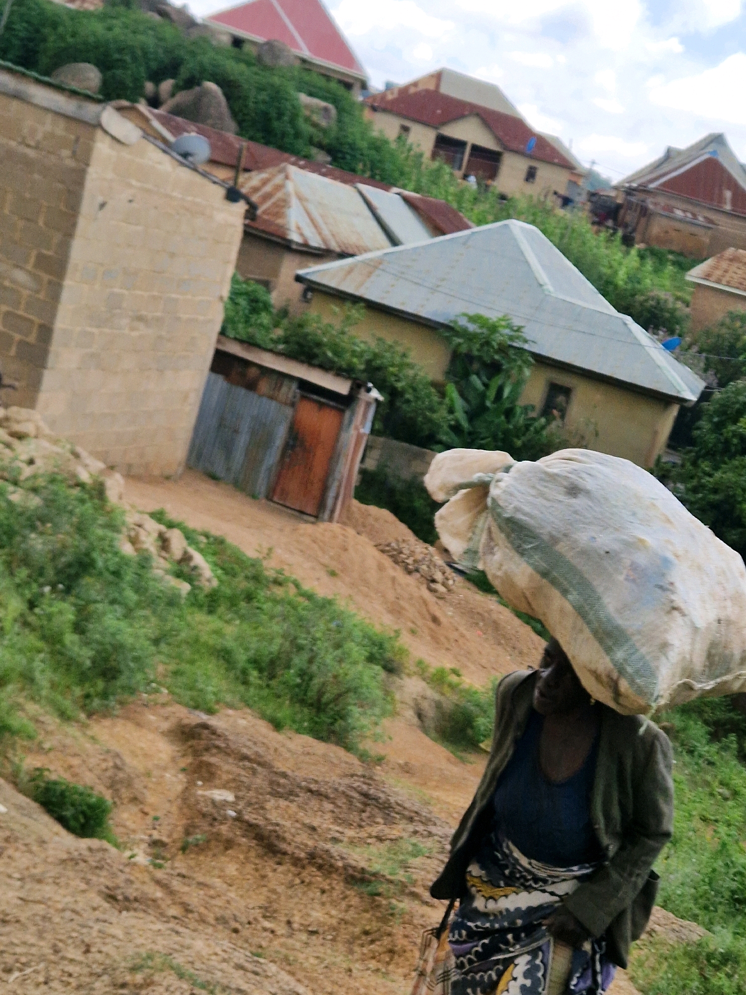 An overhead medium shot captures a woman walking uphill, carrying a large sack of produce on her head. The sack appears to be made of woven material and is tied with rope. The woman is wearing a dark blue tank top, a dark olive green jacket, and a patterned skirt. Her face is turned to the left, and she has dark skin and dark hair. She is on a dirt path surrounded by greenery and rough terrain. In the background, several houses with corrugated metal roofs are visible, situated on a hillside. The houses are constructed from brick and concrete. The sky is partly cloudy.
