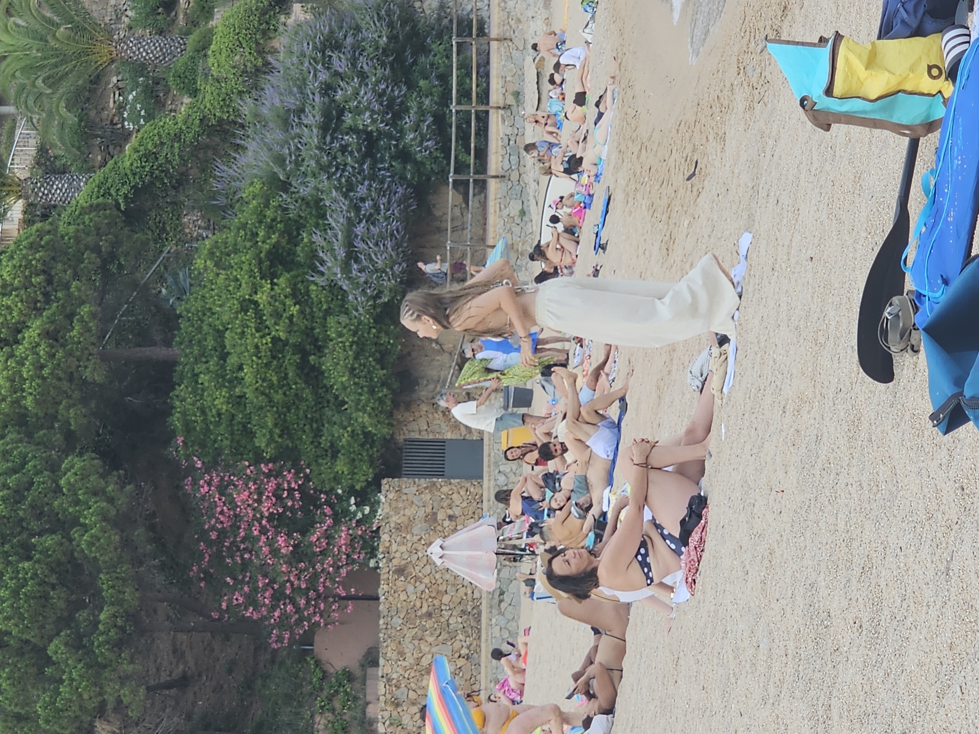 The image depicts a sunny day at a crowded beach in Blanes, Spain, likely Cala Giverola or a similar cove along the Costa Brava. In the foreground, a woman with braided hair is standing and appears to be mid-motion, perhaps dancing or posing, wearing light-colored wide-leg pants and a bikini top. Behind her, numerous people are relaxing on the sand, sunbathing, and socializing. Some are lying on towels, while others are sitting or leaning against beach bags. Beach umbrellas and towels of various colors are scattered across the sand.
The background features a lush, green hillside covered in trees and shrubs. Pink flowering bushes and purple-hued foliage are visible, adding color to the natural landscape. A stone wall runs along the base of the hill, and a wooden fence demarcates the edge of the beach area. A few palm trees are also present. The sea is visible in the far right of the image, with gentle waves lapping at the shore. The overall atmosphere is one of a popular tourist destination, with many people enjoying a leisurely day at the beach. In the lower right corner, beach equipment including a paddleboard and bags are visible.