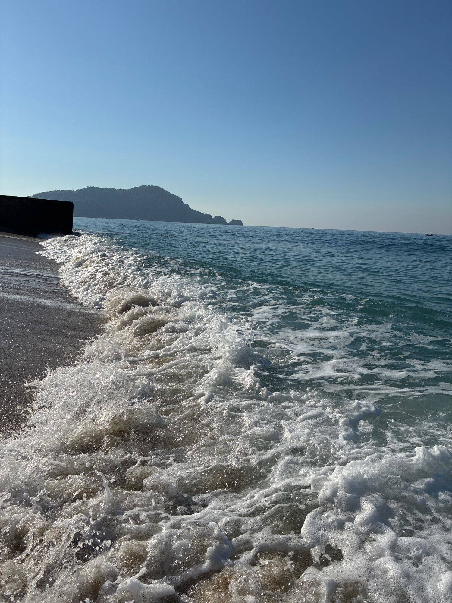 A wave crashes on the shore of Alanya, Turkey, creating a foamy white surge against the dark sand. In the background, a dark, monolithic structure, possibly a pier or breakwater, juts out into the turquoise sea. Further in the distance, a hazy, mountainous peninsula rises from the horizon, its silhouette softened by the atmospheric perspective. The sky above is a clear, brilliant blue, devoid of clouds. Two small boats are visible on the calm expanse of the sea, adding a touch of human presence to the otherwise natural scene. The sun, though not directly visible, casts a bright light, illuminating the water and creating a sense of a warm, sunny day.