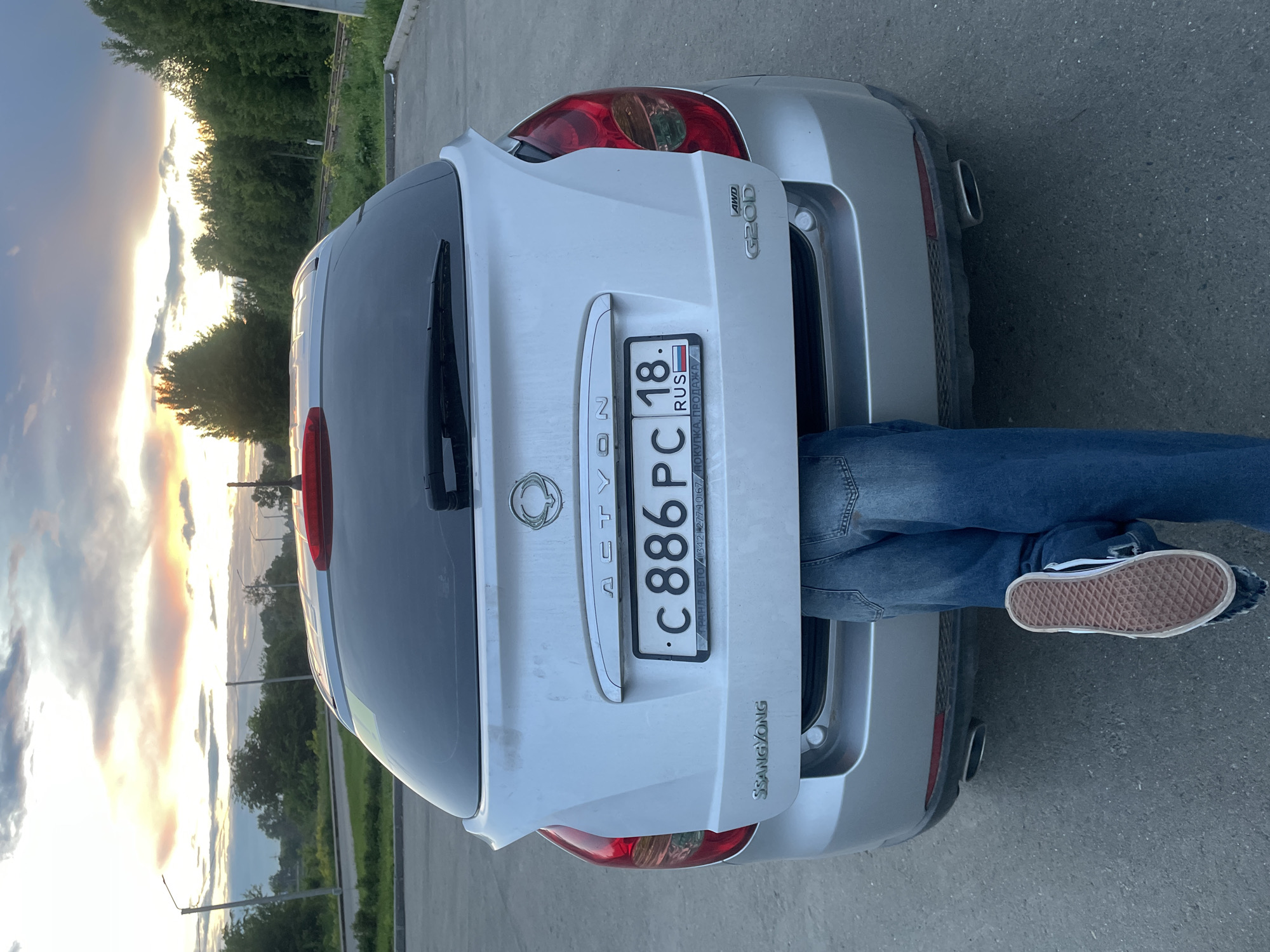 A silver SsangYong Actyon car is prominently featured from the rear, parked on an asphalt surface. A person's legs, dressed in blue jeans and light-colored sneakers with a brown sole, extend from the car's open trunk, suggesting they are partially inside the vehicle, perhaps loading or retrieving items.
The car's rear displays a Russian license plate with the registration "C 886 PC 18. RUS". Below the main number, additional text reads "ГРАНД-АВТО (342) 277 10 67 ПОКУПКА, ПРОДАЖА", which appears to be a dealership or service contact. The vehicle is clearly identified by its badging: "ACTYON" across the trunk lid, "SSANGYONG" on the lower right, and "AWD G20D" on the upper right of the tailgate. A rear window wiper and a single exhaust pipe are also visible.
The scene is set outdoors during either sunrise or sunset, characterized by the warm, glowing light on the horizon and scattered clouds, creating a dramatic sky with hues of yellow, orange, and soft purple. In the background, dense green trees line what appears to be a road or path, with glimpses of railway tracks and utility poles. The tranquil natural setting contrasts with the car and the human activity. The license plate and location context confirm the setting as Votkinsk, Russia.