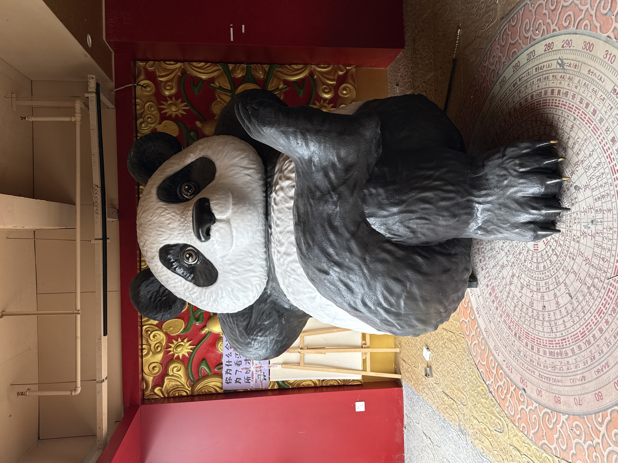 Here is a detailed description of the image:
The image shows a large, stylized sculpture of a panda bear in what appears to be a tourist or cultural setting in Yuzhong District, China. The panda is lying on its side, with its head facing the viewer. The bear's fur is depicted in a mix of white and black, with the characteristic dark patches around its eyes and ears. The sculpture looks to be made of a material like fiberglass or resin, with a texture that mimics fur.
The panda is positioned on a platform with a red and gold decorative edge. Behind the panda is a wall with intricate gold and red designs. To the right of the panda, there is a large, circular diagram featuring Chinese characters and numbers, resembling a feng shui compass or a divination chart. It appears to be set into the floor or ground.
A sign with Chinese characters is visible near the panda. A wooden easel is seen behind the panda.