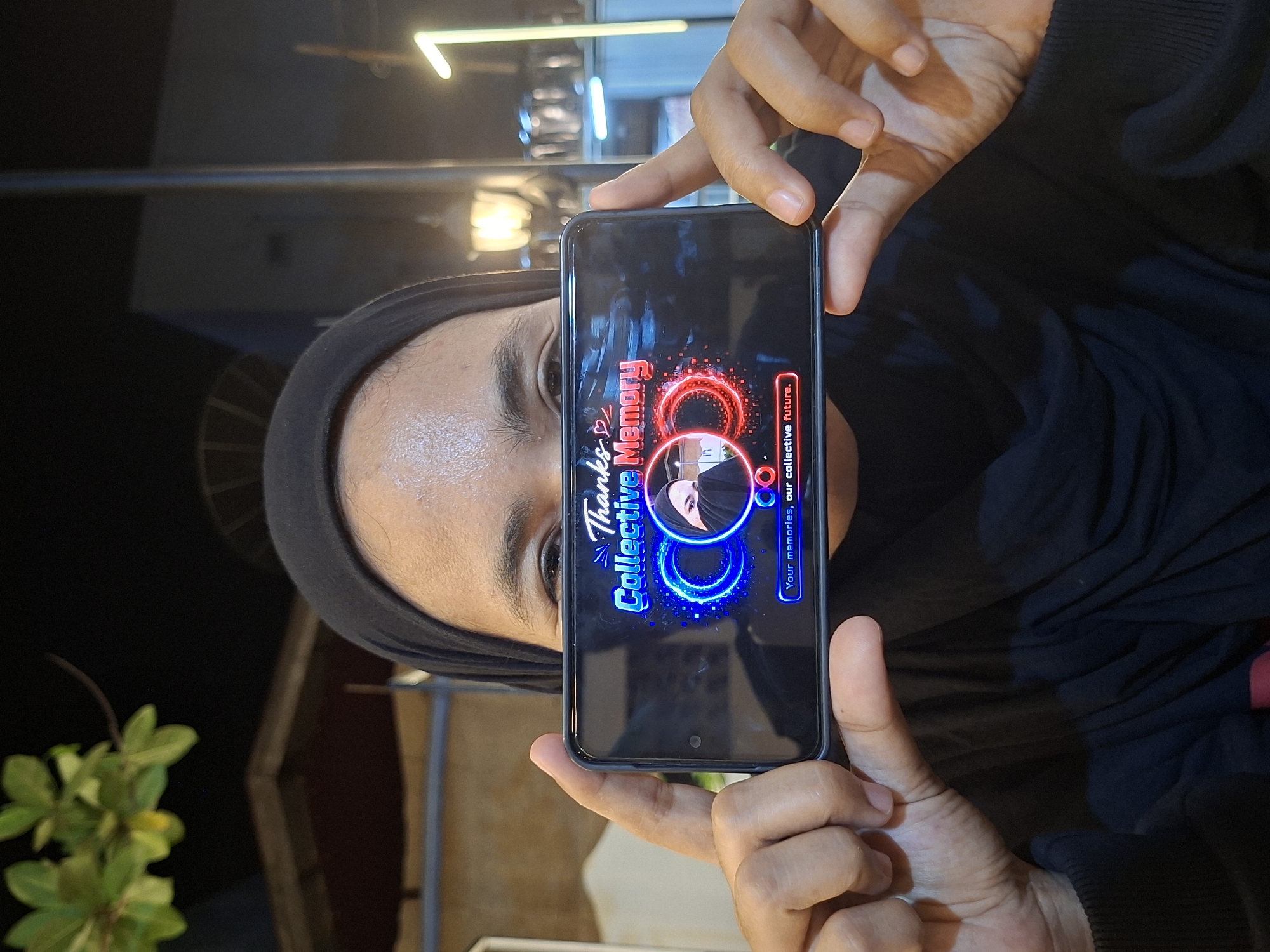 A woman in a black hijab holds up a smartphone, displaying the "Collective Memory" app interface. Her face is visible behind the phone, with direct eye contact. The phone screen shows text and graphic elements associated with the app. She is wearing a dark-colored sweater or long-sleeved shirt. Background elements suggest an outdoor setting, possibly at night. There is foliage visible in the lower left corner, and a blurred structure is visible above her head that appears to be a building with a glowing light.
Because of the information provided in the prompt, it's possible to infer that this image was taken in Lhokseumawe, Indonesia.