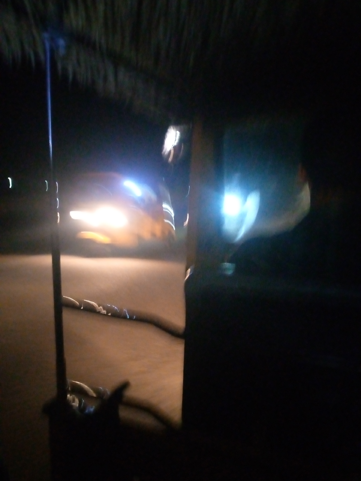 The image depicts a dark, blurry scene at night, likely in Jalingo, Nigeria. A bright headlight from a vehicle illuminates the foreground, casting a yellow and white glow on a paved surface. The light streaks suggest motion blur, indicating the vehicle is either moving or the camera is panning. A dark, vertical pole is present in the left foreground, possibly a street light or part of a structure. In the background, a dimly lit, indistinct object with a bright, scattered light source is visible, and what appears to be a thatched roof or canopy hangs overhead. The overall impression is of a low-light, possibly urban environment, with a focus on the interaction of artificial light with the surroundings.