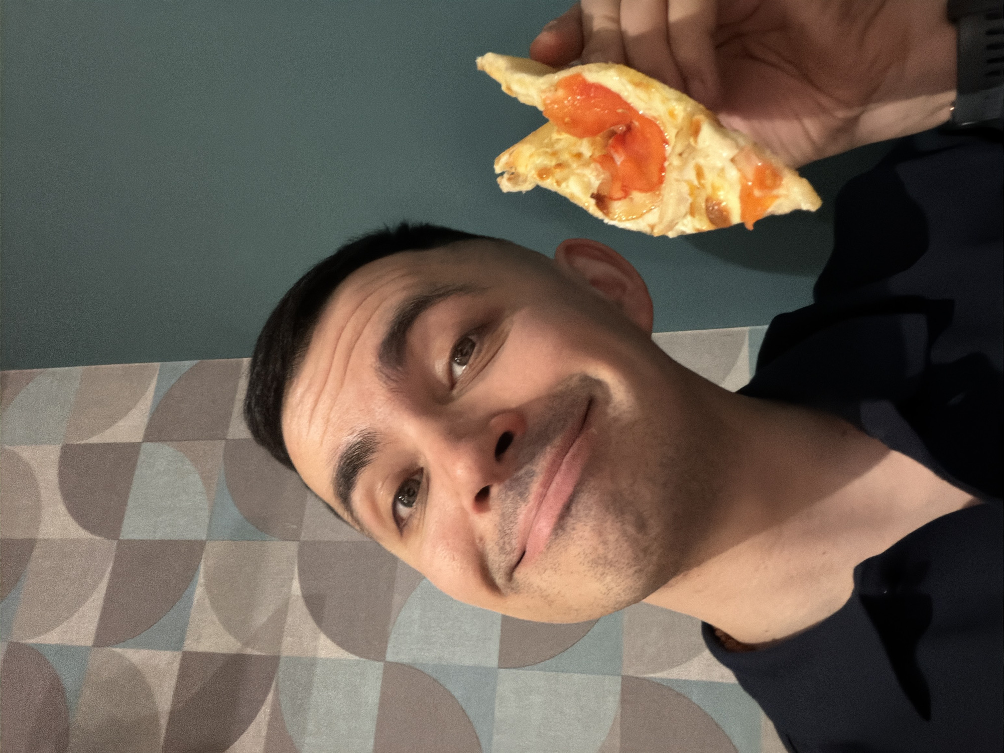 A young man is depicted indoors in Izhevsk, Russia, looking directly at the camera with a subtle smile and a slightly amused expression. He holds up a slice of pizza topped with visible red tomatoes and melted cheese, positioned in the upper right portion of the frame, suggesting an imminent meal or a pose with food.
The background features a plain dark teal wall on the upper left and a patterned wall or surface with geometric circular and triangular designs in shades of grey and light blue on the lower right. The man has short, dark hair and appears to be wearing a dark-colored shirt. A dark watch is partially visible on his right wrist. The specific time of day is not apparent from the indoor lighting. No text is visible in the image.