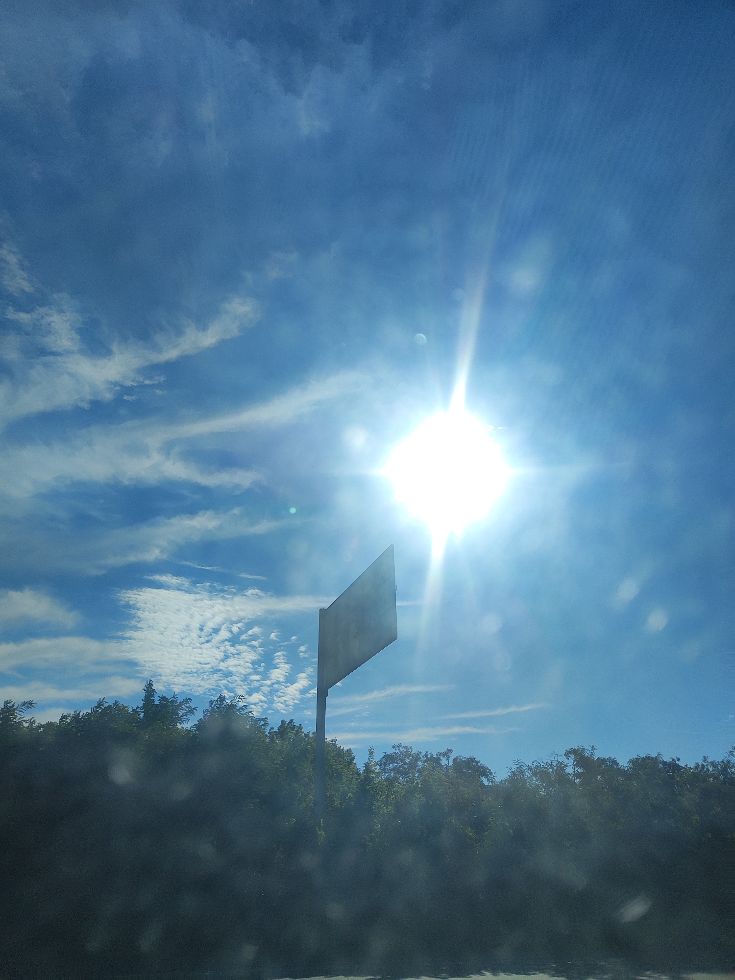 A bright sun shines in a bright blue sky with scattered white clouds. A tall, dark pole stands in the center of the frame, topped with a square, angled sign. The sign is tilted to the left, and its surface appears weathered and faded. The background is filled with the tops of lush green trees, their leaves dark against the bright sky. The bottom portion of the image is obscured by a dark, blurry foreground, possibly a car window.