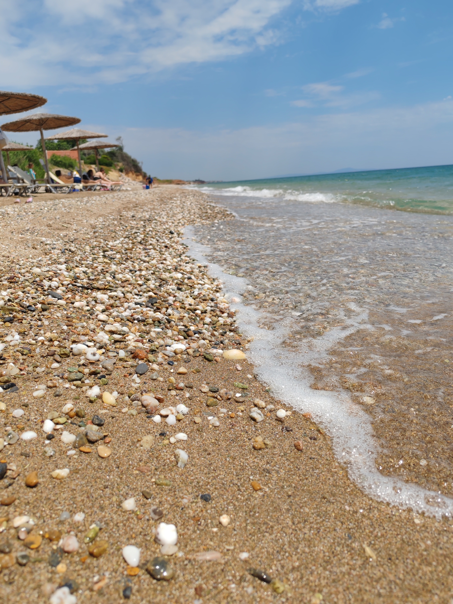This is a low-angle, close-up, eye-level shot of a pebble and sand beach in Makri, Greece. The foreground is dominated by wet pebbles and sand, with the edge of the clear turquoise sea lapping at the shore, creating a line of white foam. In the mid-ground, several thatched umbrellas and lounge chairs are visible, with a few blurry figures relaxing on them. The background shows a sandy cliff face, more umbrellas, and the expansive blue sky with scattered white clouds. The sea stretches out to the horizon, where a faint landmass is visible. The overall impression is a peaceful and sunny day at the beach.