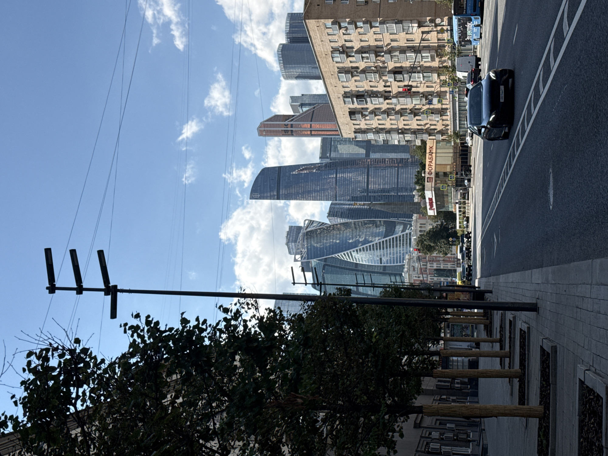 The image captures a street scene in Moscow, Russia, with the Moscow International Business Center (MIBC), also known as Moscow City, as the prominent backdrop. The foreground features a tree-lined sidewalk with newly planted trees, their trunks wrapped in protective material. A dark-colored sedan is in the right lane of a multi-lane road. Beyond the road, a low-rise building with multiple windows and balconies is visible, with a traffic light at an intersection. Further in the background, the iconic skyscrapers of Moscow City dominate the skyline under a bright, partly cloudy sky. One of the prominent skyscrapers is the Neva Towers, recognizable by its twisted glass facade. A light blue bus with Russian text, possibly an advertisement, is also present on the road. The overall scene depicts a modern urban environment in Moscow.