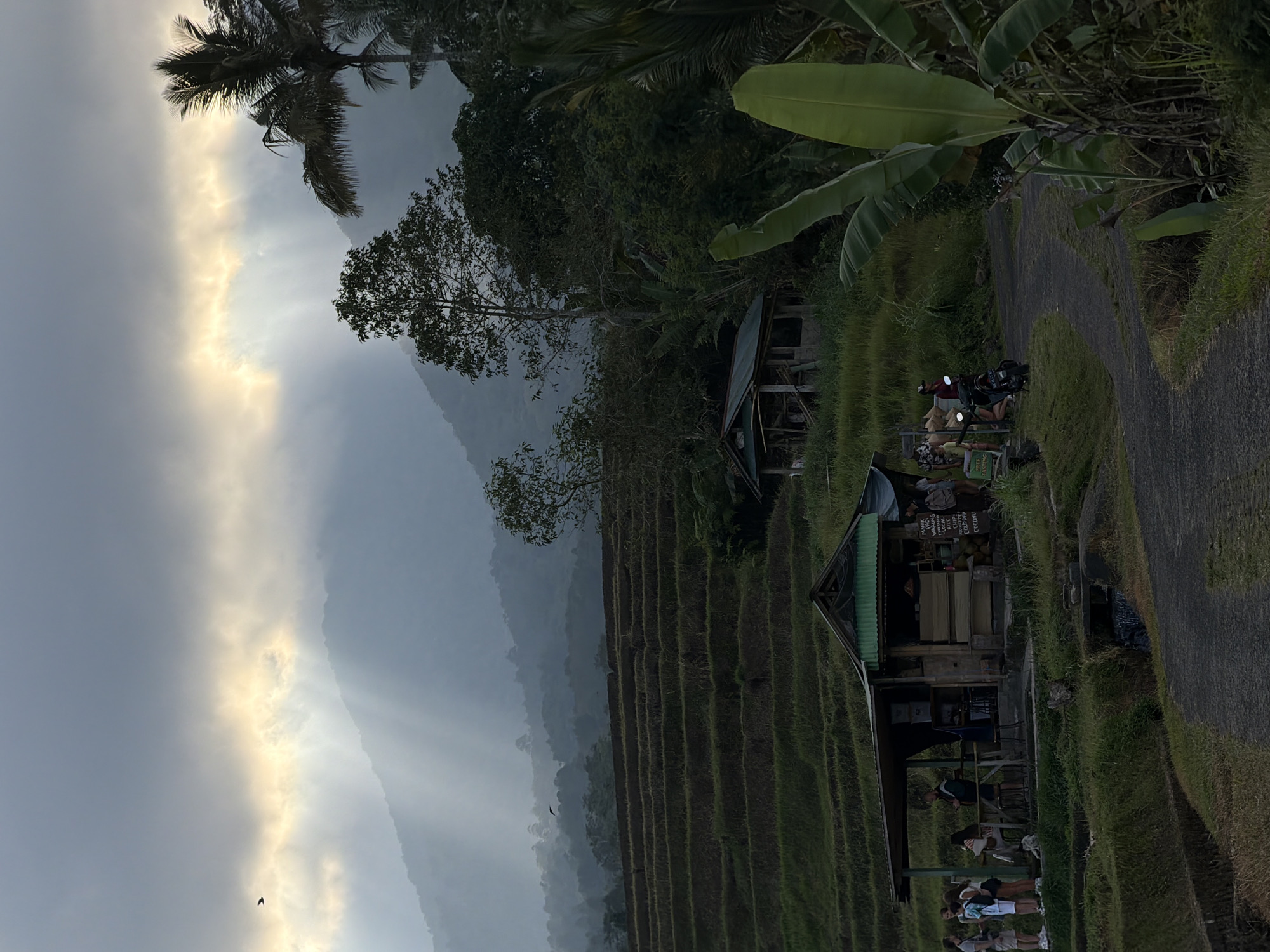 This image depicts a scenic view of the rice terraces in Penebel, Indonesia, likely during the early morning or late afternoon due to the soft, diffused lighting. The sky is dramatic with rays of light breaking through clouds, illuminating the misty mountains in the background.
In the mid-ground, a small roadside stall is visible, with several people standing around it, possibly tourists. The stall appears to sell refreshments or local goods, with signs written in Indonesian. A scooter is parked near the stall, and a person is riding another scooter away from the scene on a paved path.
The rice terraces themselves dominate the left side of the image, showcasing the characteristic tiered cultivation. Lush green vegetation, including banana plants, adds to the tropical atmosphere. A small, rustic structure, possibly a hut or storage building, is situated amidst the greenery. The overall impression is one of tranquility and natural beauty, characteristic of rural Bali.