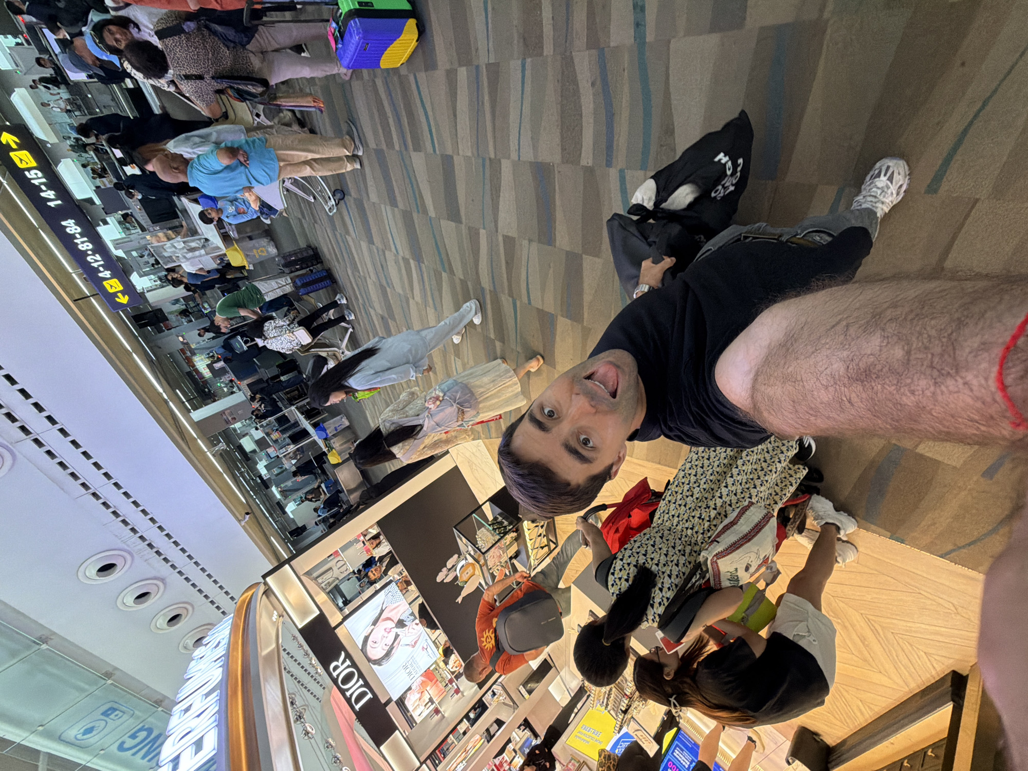 The image shows a man taking a selfie at an airport in Tambon Mai Khao, Thailand, near a Dior store. He is wearing a black t-shirt, jeans, and white sneakers. Other people are in the vicinity, some waiting in line at desks while others are shopping. Luggage is present. Signage indicates gate numbers 14-15 and 4-12-81-84. The flooring is patterned with neutral colors.