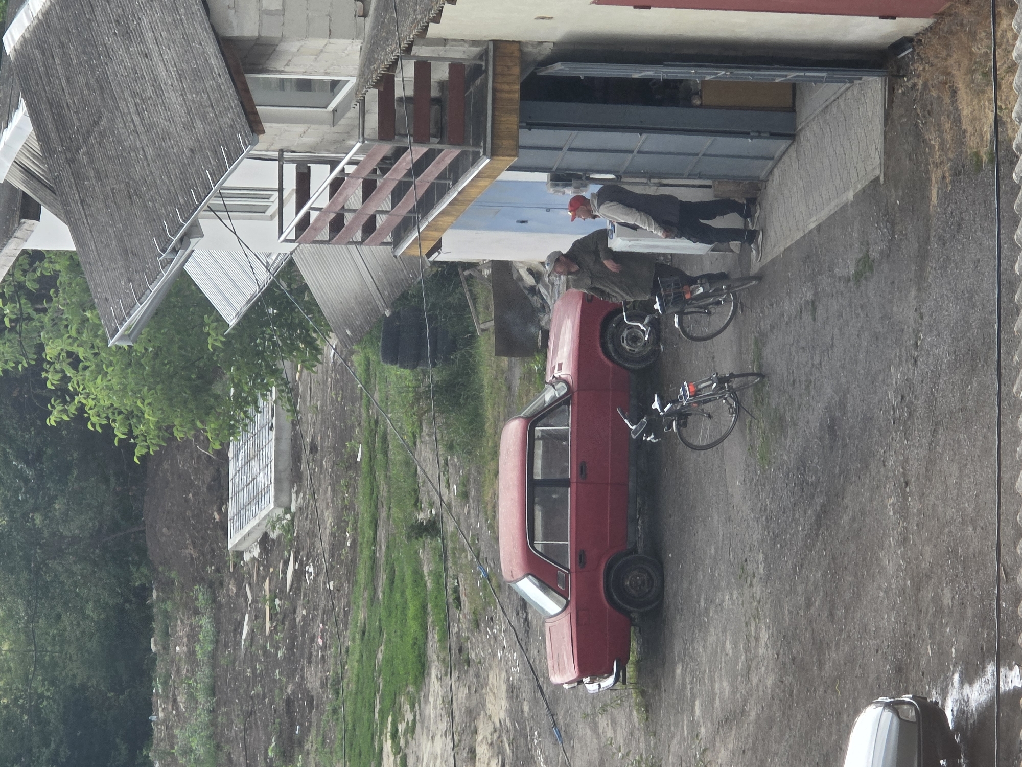 The image shows an outdoor scene in Dubno, Ukraine, with a red sedan parked on a dirt ground. Two men are standing near the entrance of a building, one wearing a green jacket and a cap, and the other in a vest and a red cap. Several bicycles are also present in the scene, including one leaning against the car and another one parked nearby. The building appears to be a workshop or garage with a metal door and a wooden staircase leading to a balcony. The surrounding area is somewhat unkempt, with some debris and vegetation visible. In the background, there is a hill with trees and a concrete structure.