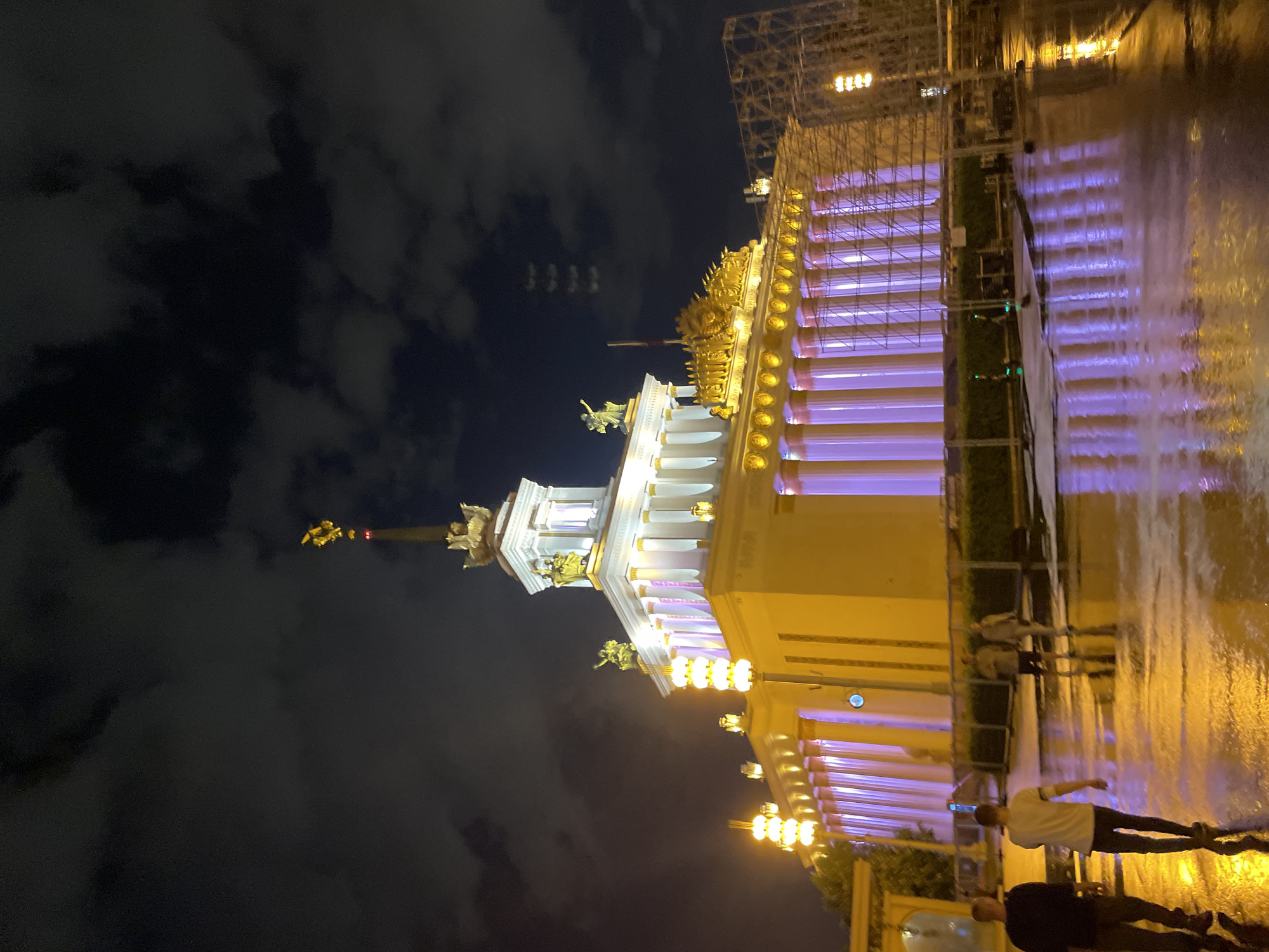 The image depicts the illuminated pavilion #1 "Central" at the VDNKh exhibition park in Moscow, Russia, at night. The structure is brightly lit with white and golden lights, with purple-lit pillars. The sky is dark with clouds. The foreground is wet and reflects the lights. Two people are visible walking across the wet surface in the lower right. Scaffolding is visible on the right side of the building.