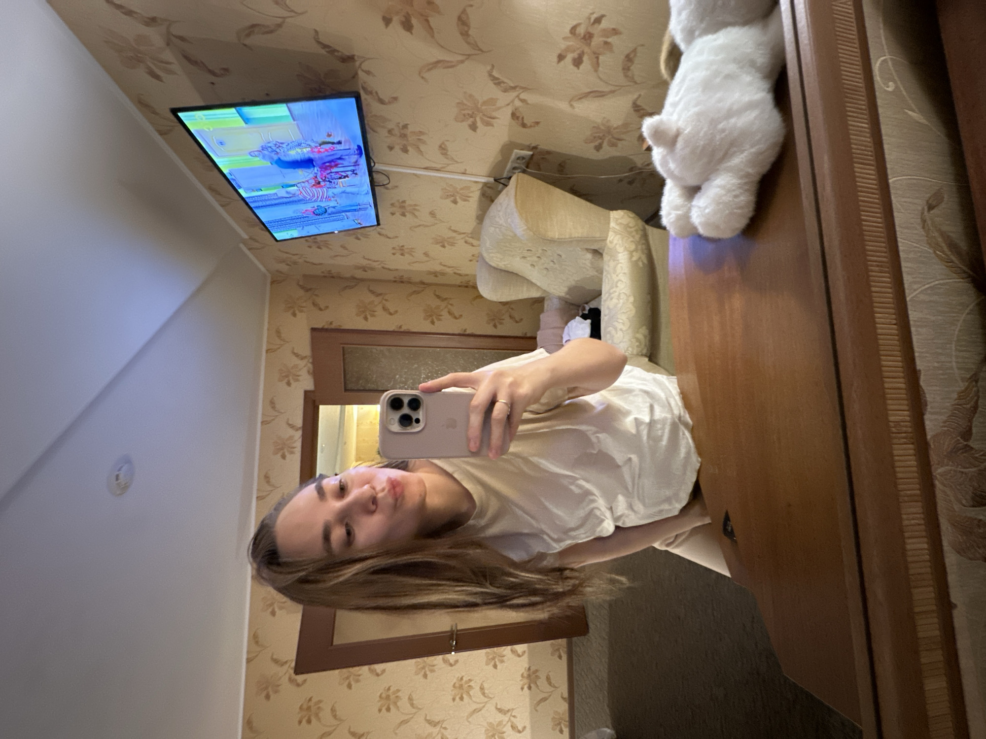 A young woman stands inside an apartment in Yekaterinburg, Russia, taking a selfie. The room features patterned wallpaper, a television mounted on the wall, a chair, and a wooden cabinet with a plush toy on top. The woman is wearing a white shirt and holding a smartphone.