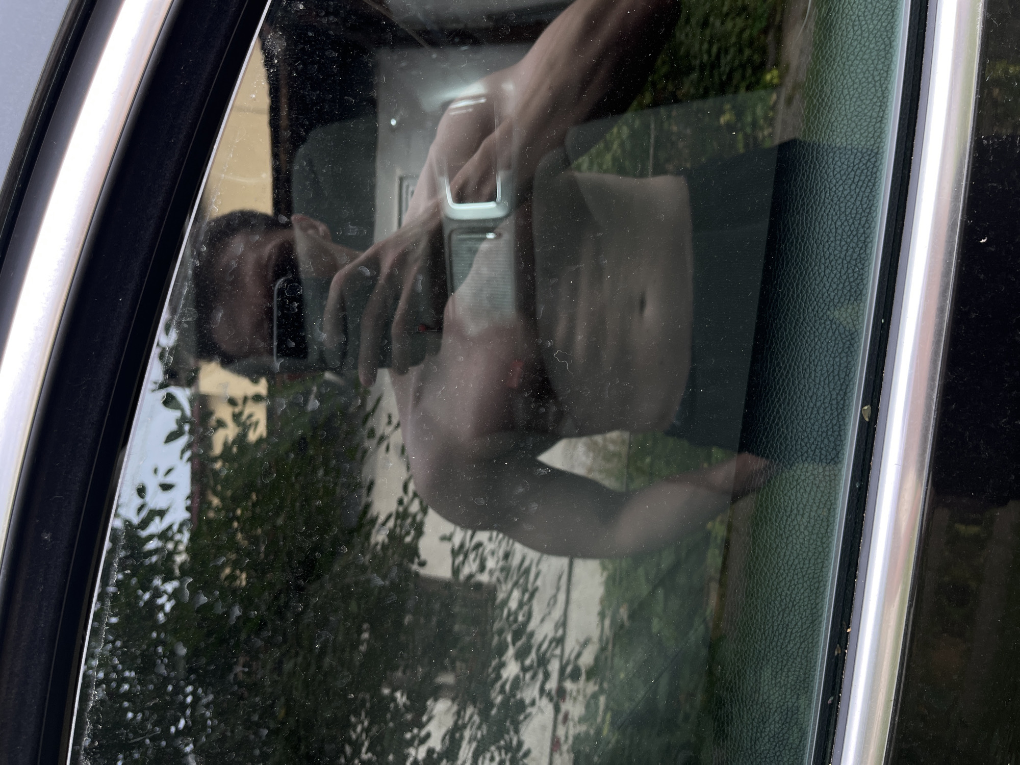 The media file is a photograph taken from the side of a car, showing a reflection in the window. The reflection captures a shirtless person holding a smartphone, presumably taking a selfie. The person's torso and arms are visible, with a muscular build. The background of the reflection appears to be an outdoor setting with greenery and possibly buildings, suggesting a daytime scene. The car window is somewhat dirty, with specks of dust and water spots visible, which may distort the reflection slightly. The photograph also shows the interior of the car's door frame, with textured material and a metallic trim. There is no visible text in the image. The overall impression is a candid moment captured by a reflection in a car window.