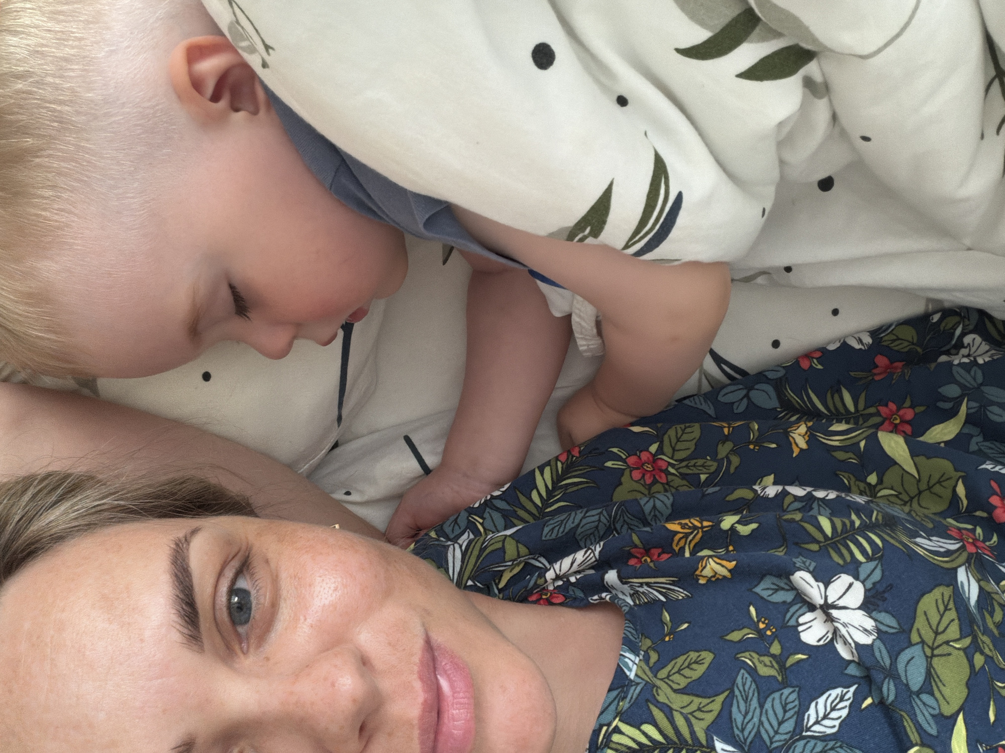 A woman with light hair and blue eyes lies in bed, wearing a blue floral-patterned shirt. A young child with blond hair lies next to her, partially covered by a white blanket with black dots and green leaf designs. The child appears to be looking at the woman. They are lying on a white bedsheet with faint gray stripes. The location is not discernible in the image, so the city of Votkinsk, Russia cannot be confirmed.