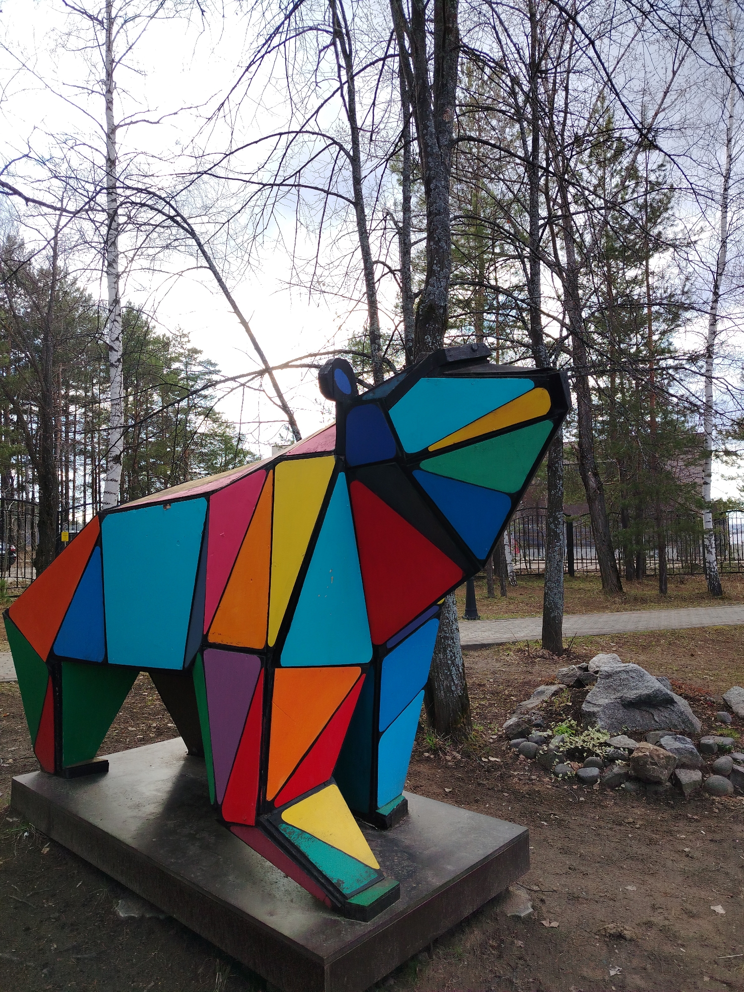 A vibrant, polygonal sculpture of a bear stands as the central subject in what appears to be a park or forested area in Chaikovsky, Russia. The bear is crafted from numerous flat, geometric panels, each painted a distinct bright color such as red, blue, green, yellow, orange, purple, and cyan, with black outlines separating the segments. The sculpture is posed in a walking or sniffing stance, with its head slightly elevated, and rests on a dark, rectangular base.
The setting is characterized by a mix of trees, primarily bare deciduous trees with visible white-barked birches, intermingled with evergreen coniferous trees like pines, suggesting either late autumn or early spring. A paved pathway winds through the trees in the mid-ground, bordered by a black metal fence. A building with a dark roof and lighter walls is faintly visible in the far background.
The ground around the sculpture is earthy, with patches of sparse grass and some fallen organic matter. To the right, a small arrangement of rocks and stones, interspersed with subtle green vegetation, adds to the natural feel of the immediate foreground. The sky is bright and overcast, creating soft, diffused lighting across the scene without casting strong shadows. There are no people or discernible activities taking place, contributing to a serene and contemplative atmosphere. No visible text is present in the image.
