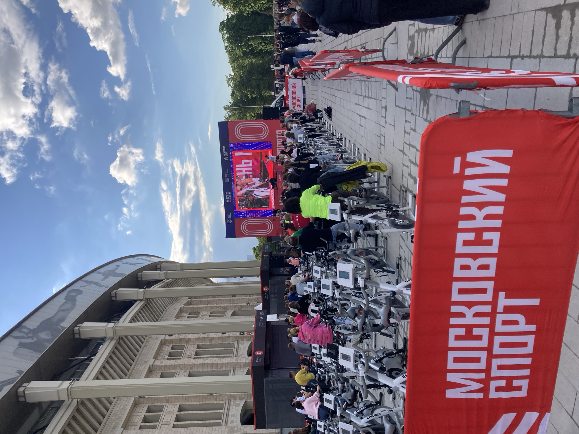 The image shows an outdoor event taking place in Moscow, Russia. In front of a building with a distinctive curved roof are rows of stationary bikes. People are seated on the bikes. A large screen displays content for the audience. Red banners with white Cyrillic text are present. Banners with white stripes are also visible. The sky is blue with clouds. The phrase "Московский Спорт" translates to "Moscow Sport," indicating the event is related to sports in Moscow.