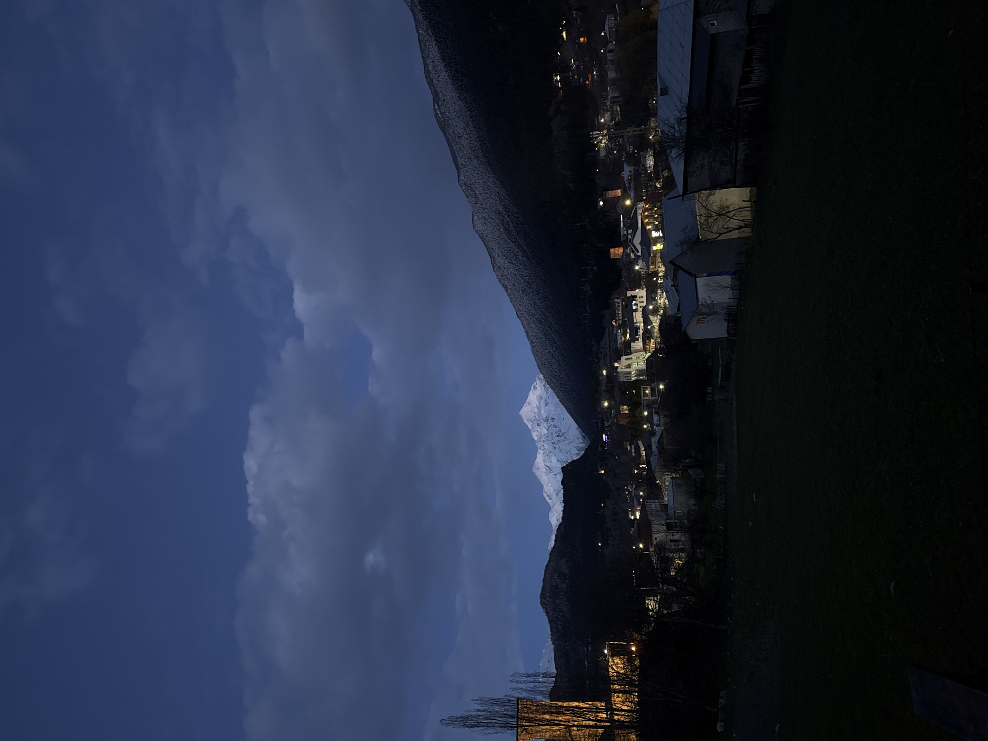 The image depicts a nighttime scene in Mestia, Georgia. A snowy mountain peak is visible, lit by moonlight or artificial light. The town's lights illuminate buildings and streets below. The sky is cloudy with varying shades of blue. The foreground consists of dark areas, possibly fields or foliage. A building with illuminated windows is also visible.
