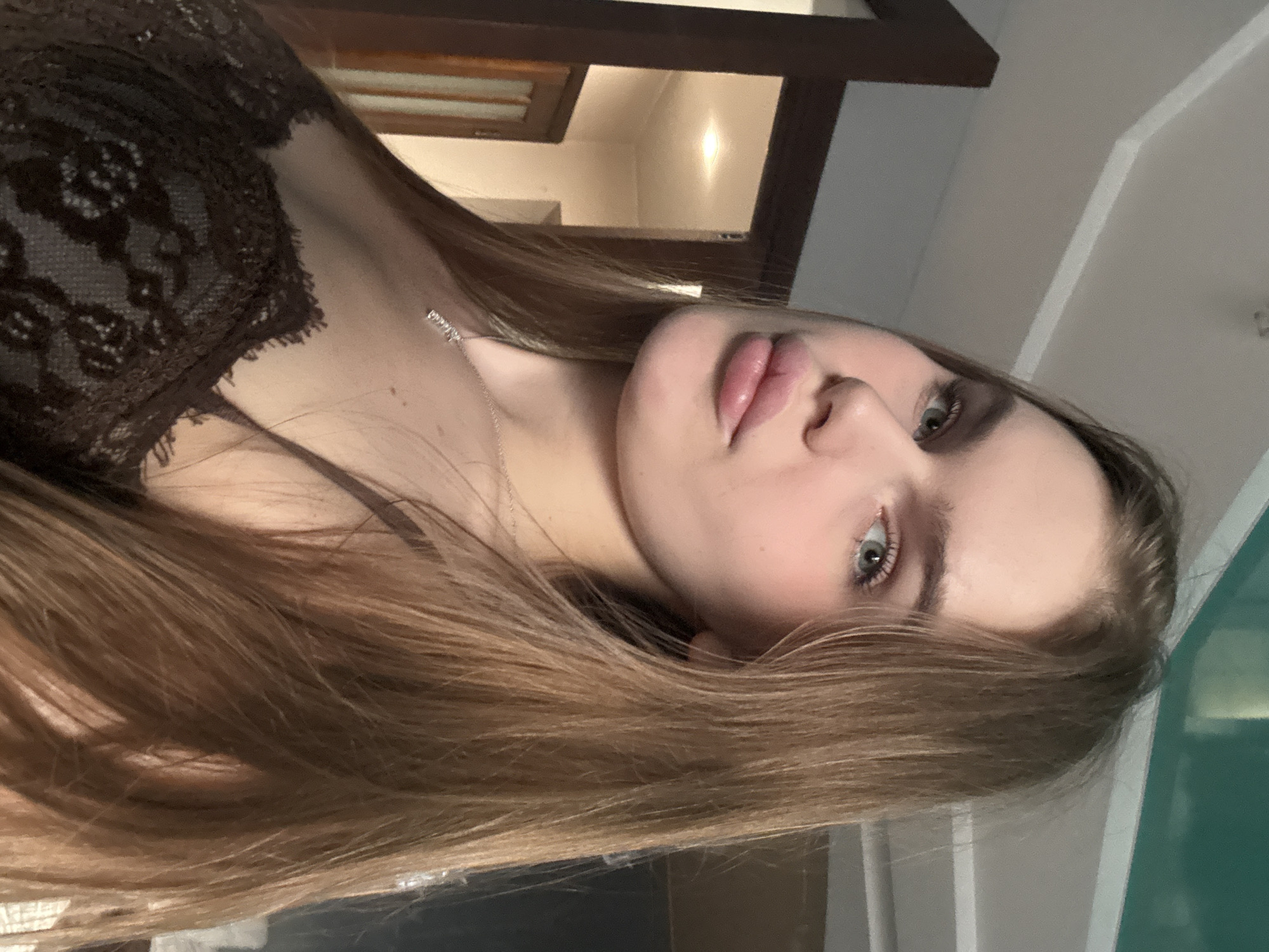 A young woman is pictured in a close-up, sideways shot. She has long, light brown hair and fair skin. Her lips are plump and slightly parted, and she is wearing a delicate silver necklace. She appears to be wearing a dark lace top. The background is out of focus but suggests an indoor setting with wooden trim and light-colored walls, possibly a room in a house. The lighting is soft, suggesting it could be daytime or artificial lighting. The woman's gaze is directed slightly away from the camera, and her expression is neutral. There is no visible text in the image. The location context is Dnipro, Ukraine.